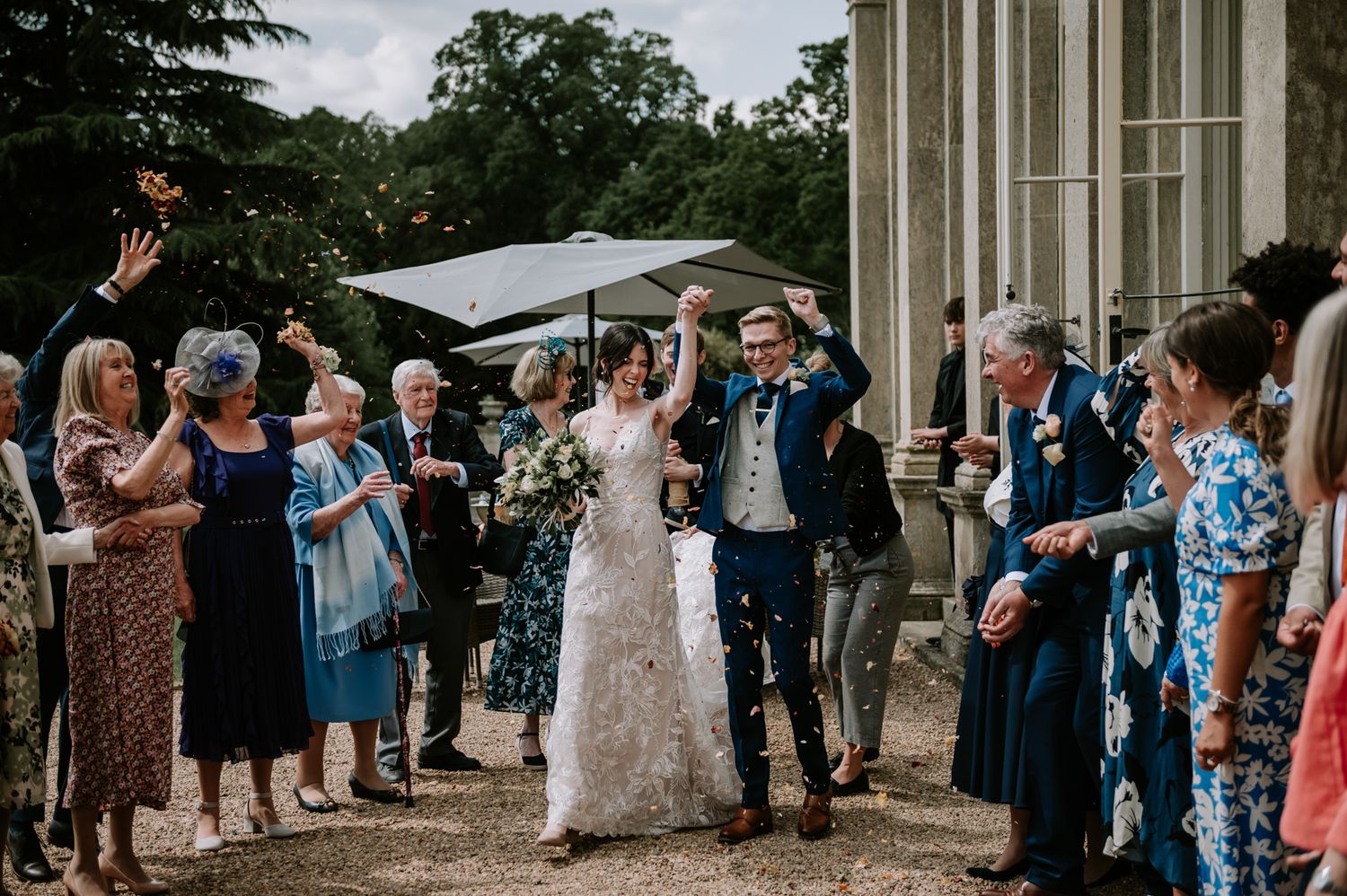 Newlyweds walk together through a quaint village courtyard surrounded by well-wishers and historic brick buildings.