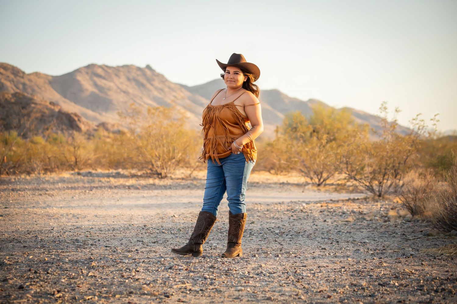 Cowboy in a brown leather jacket walks through scenic desert landscape during golden hour.
