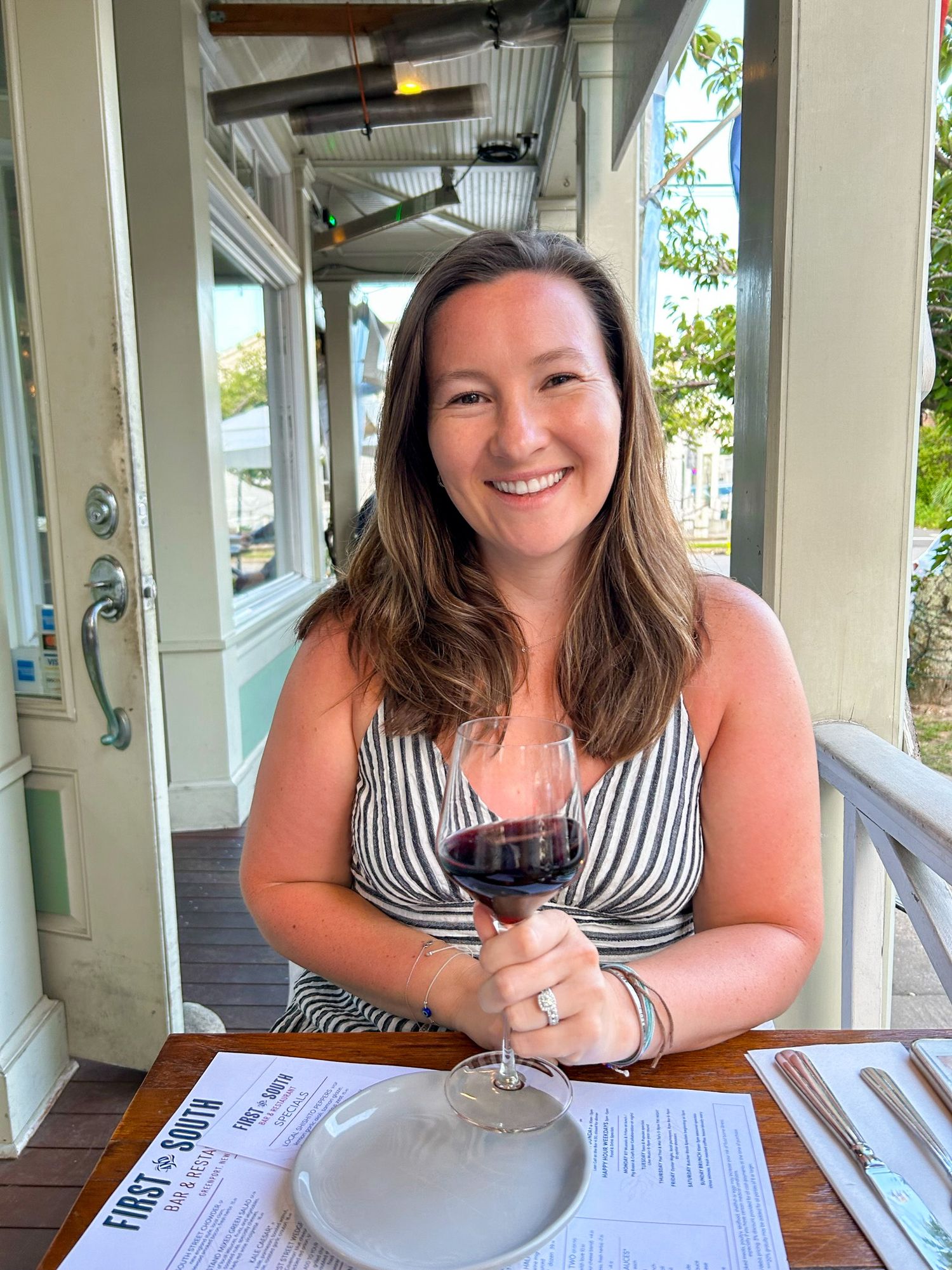 Woman in striped dress sitting at outdoor restaurant table with menu and place settings.