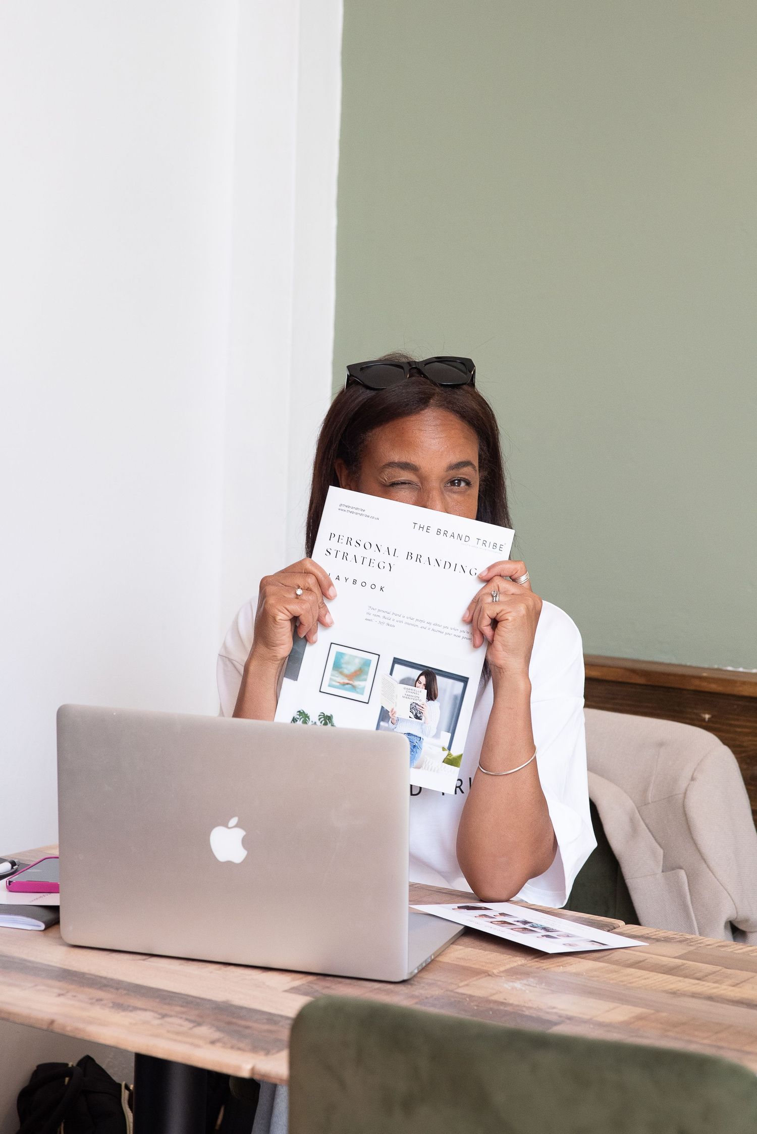 Someone sits at a wooden desk with a MacBook laptop and holds up a paper document in front of their face.