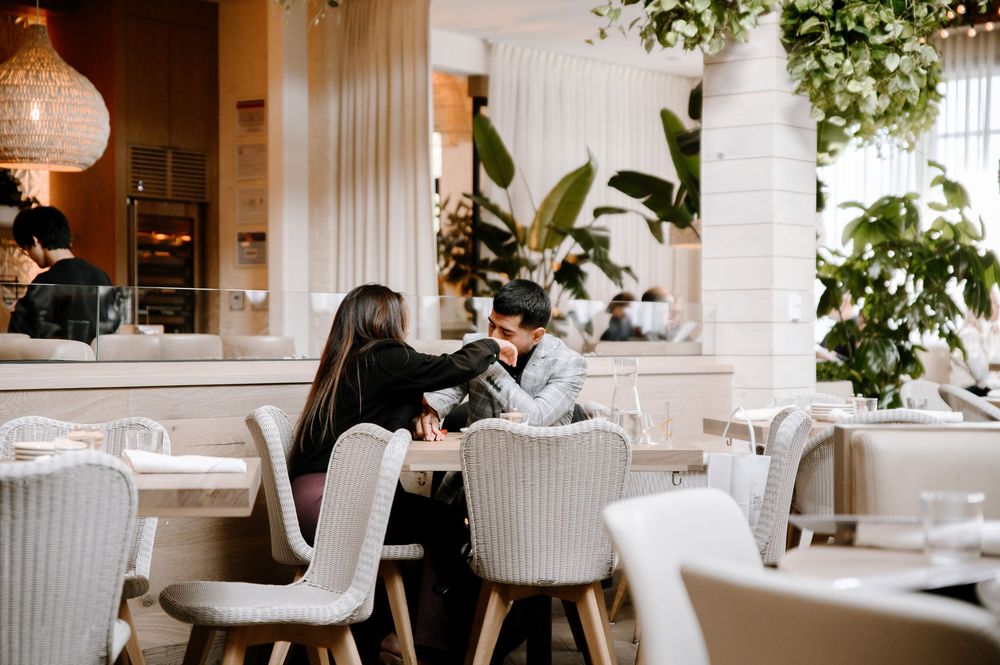 Interior of a bright modern restaurant with white decor, plush chairs, and lush green plants throughout.
