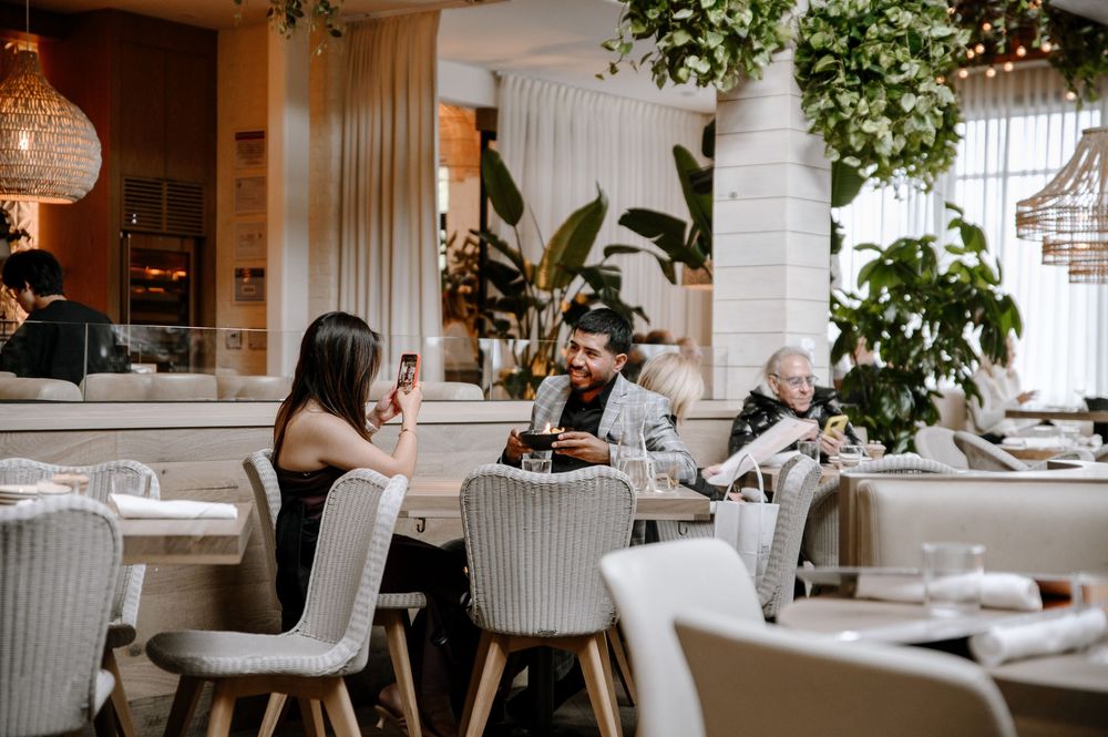 Modern restaurant interior with patrons dining at tables decorated with hanging lights and greenery.