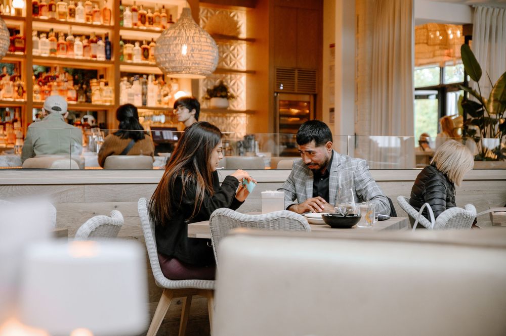 Diners seated at elegant white tables in an upscale restaurant with warm lighting and shelves of bottles behind the bar.