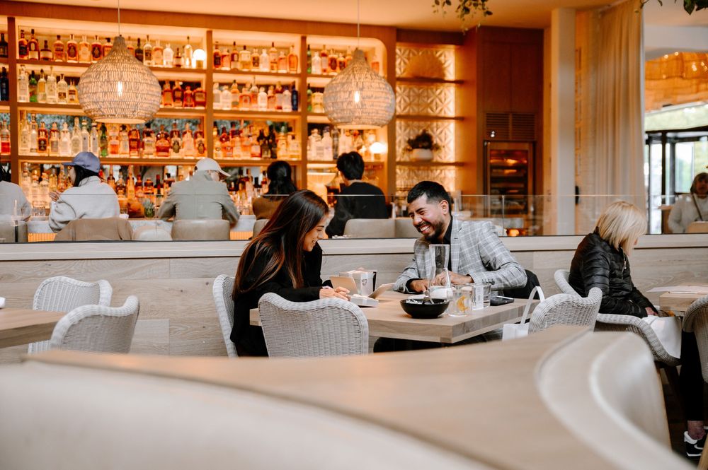 Diners enjoying drinks and conversation at an upscale restaurant bar with warm lighting and wooden shelving.