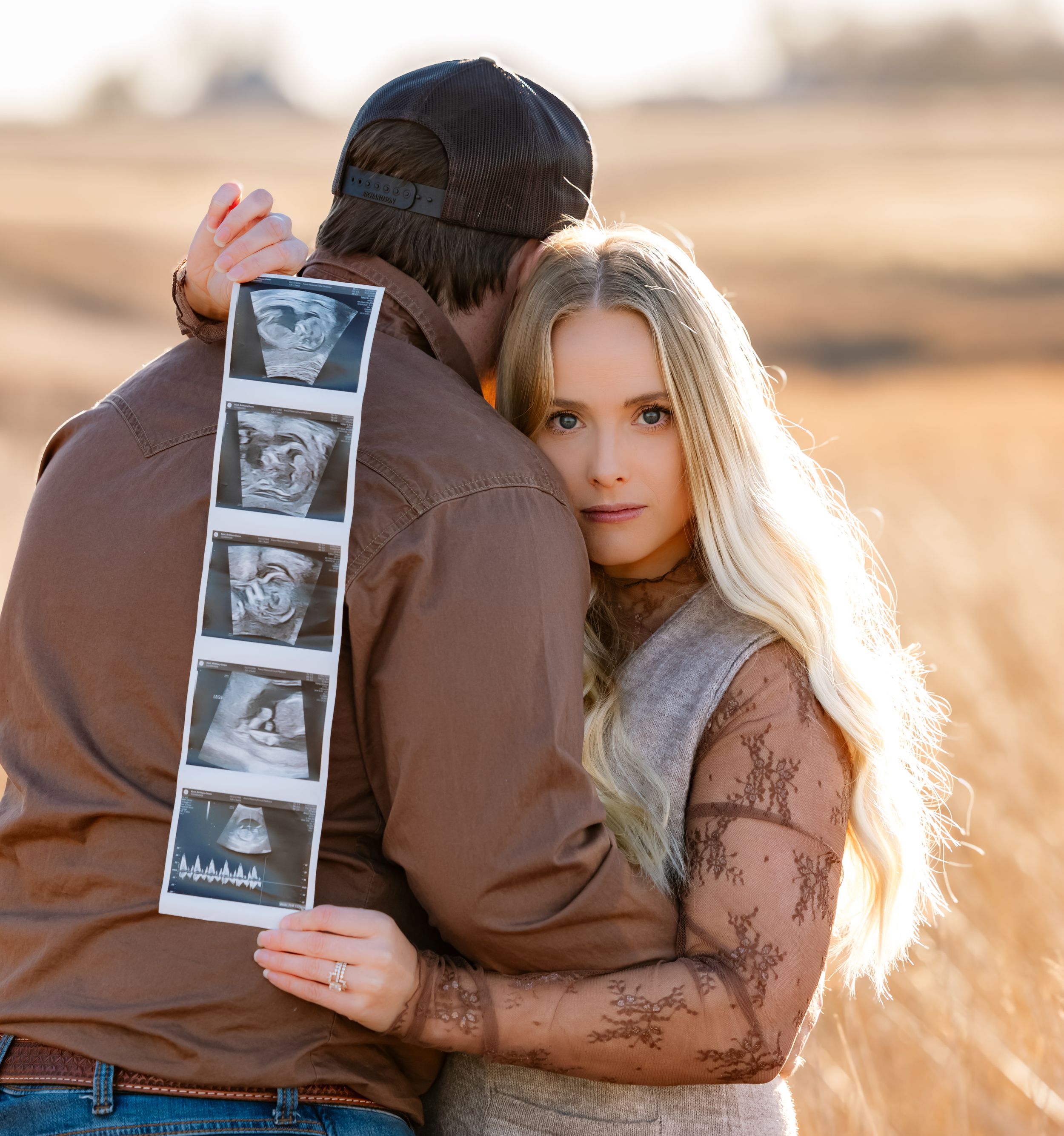 A couple holds up a strip of ultrasound photos in a sunlit field during a pregnancy announcement photoshoot.