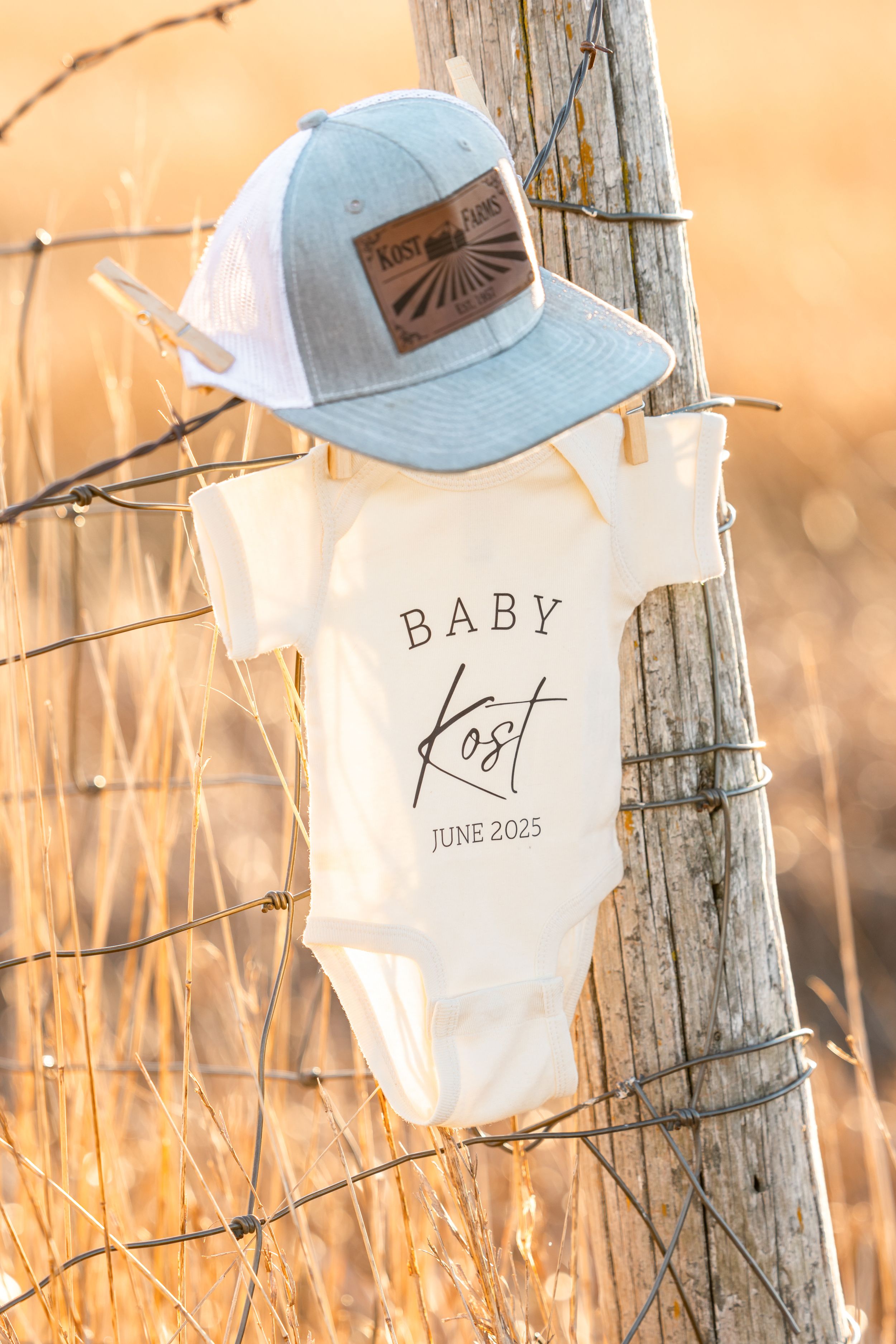 Baby announcement display with onesie and baseball cap hanging on rustic fence at sunset.