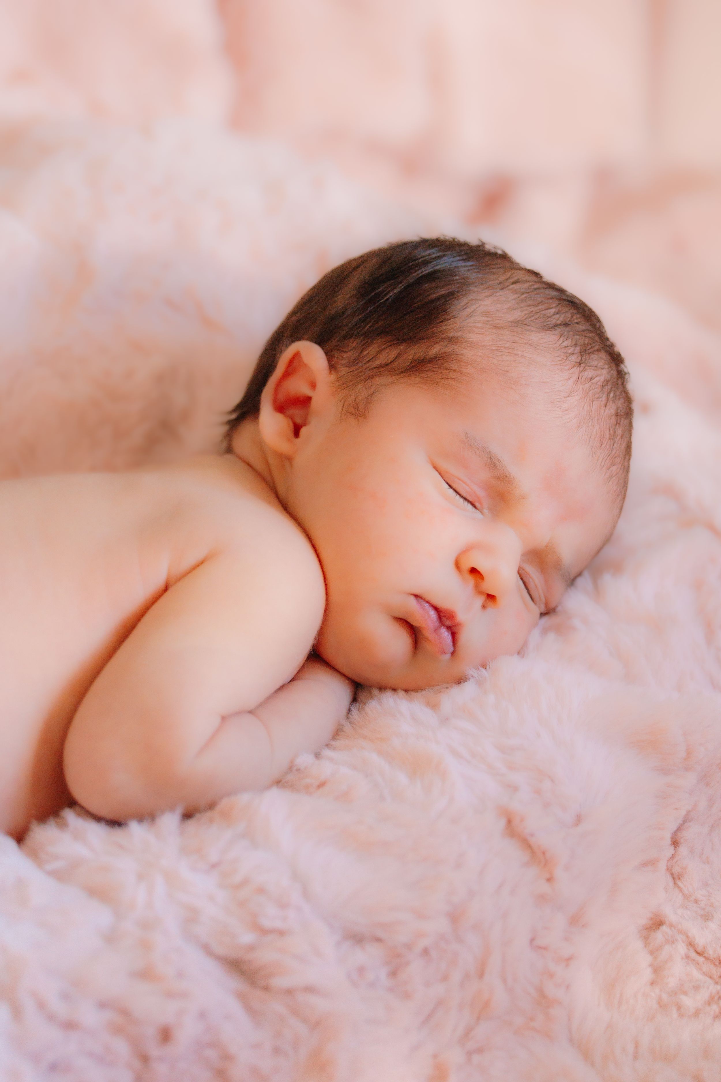 Peaceful newborn baby sleeping soundly on soft pink fuzzy blanket in natural lighting.