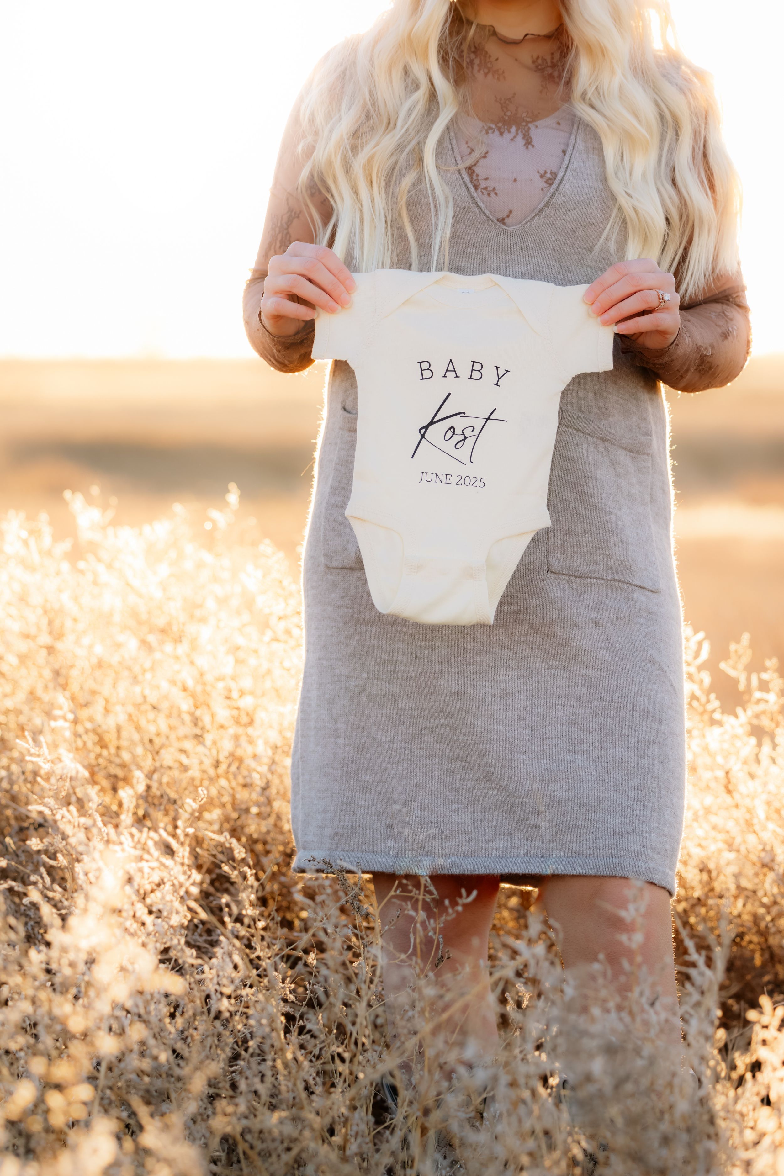 Someone holds up a white baby onesie in a golden field during sunset for a pregnancy announcement photo.