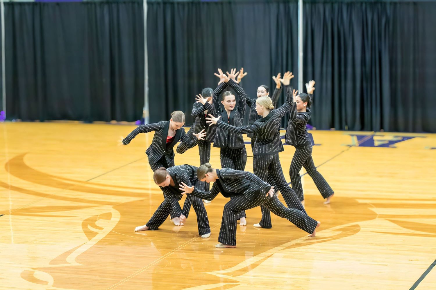 Dance team performs synchronized routine wearing all black outfits on yellow gymnasium floor during competition.