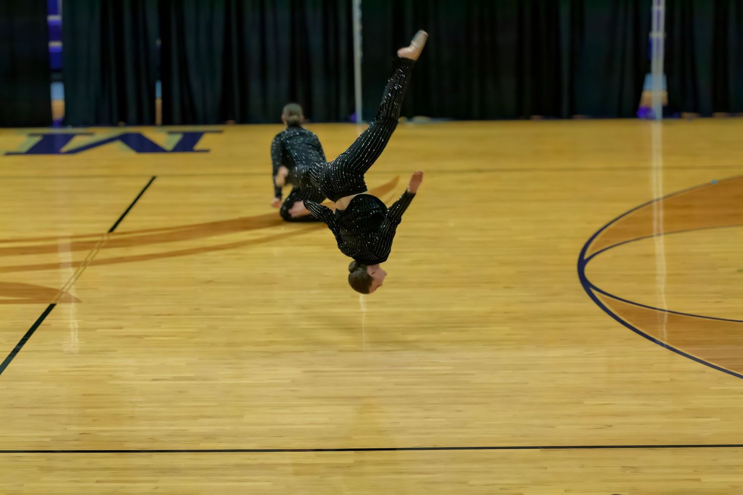 Break dancer performing a complex headstand move with legs extended in the air on a polished gym floor.