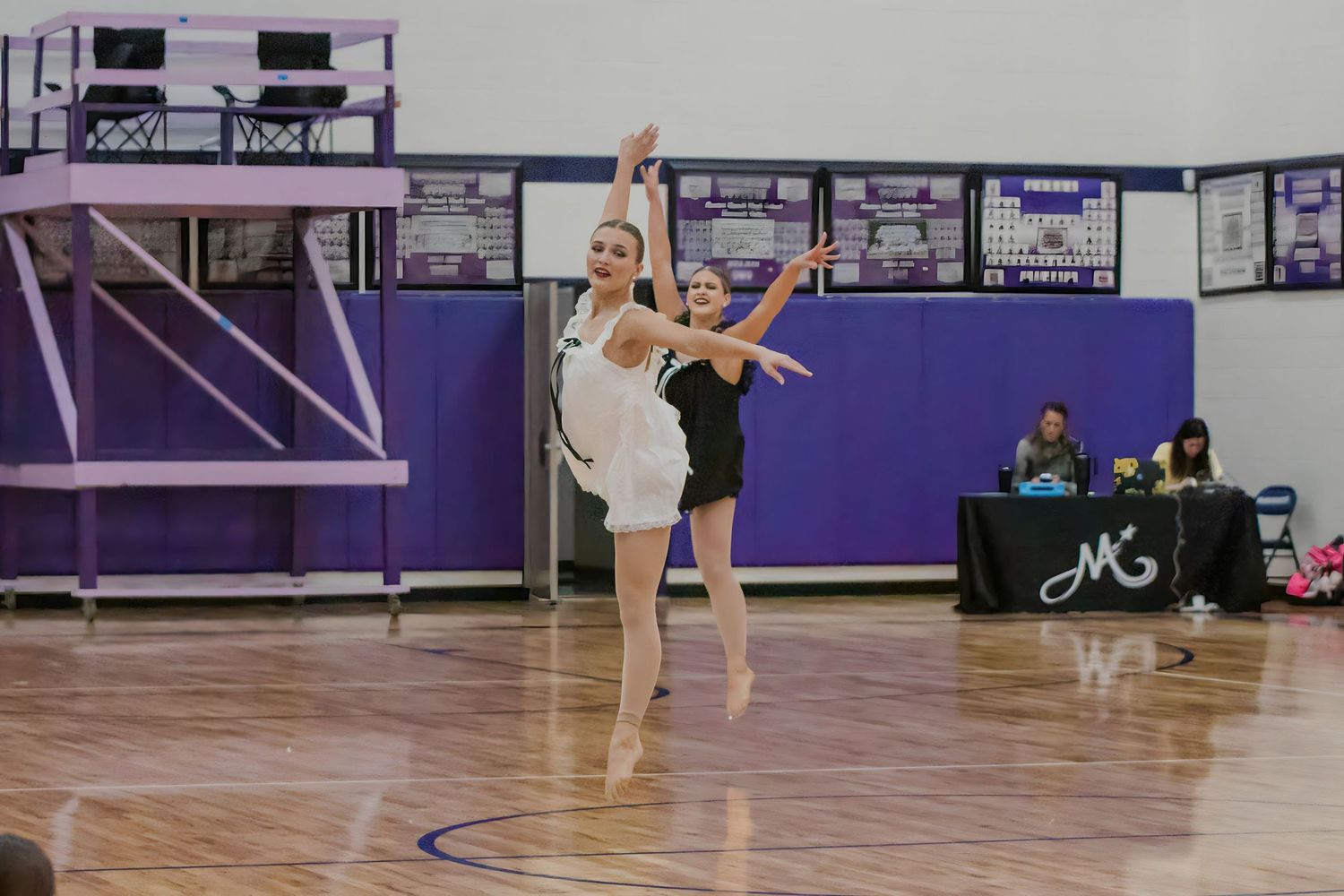 Volleyball players in action on a gymnasium court with purple walls and athletic equipment visible in the background.