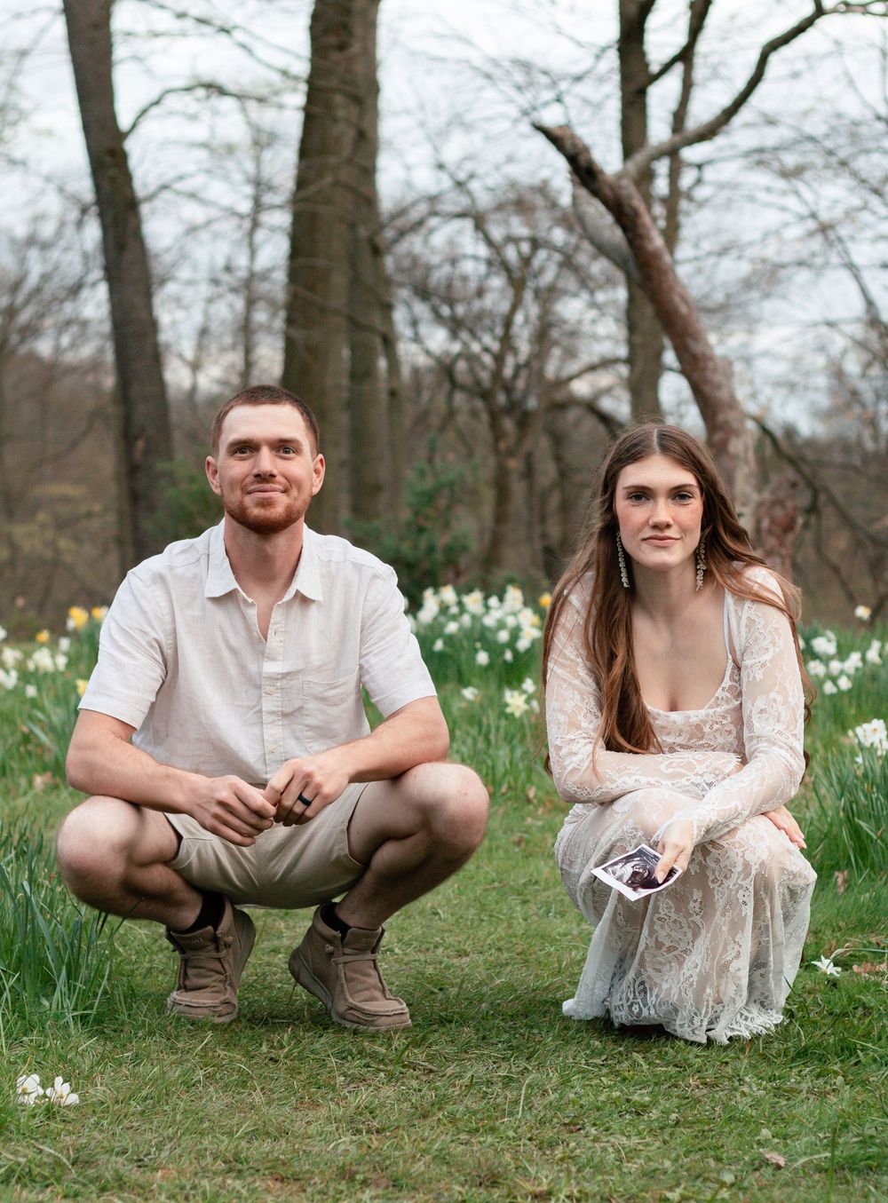 A couple poses together in a spring meadow filled with blooming white daffodils among bare trees.