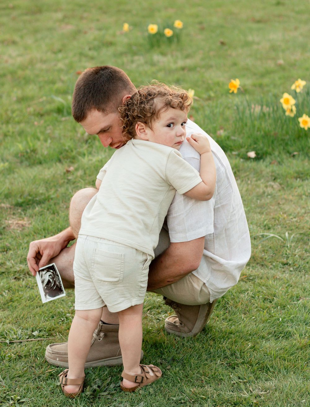 Two people share a tender moment in a grassy field with yellow daffodils blooming in the background.