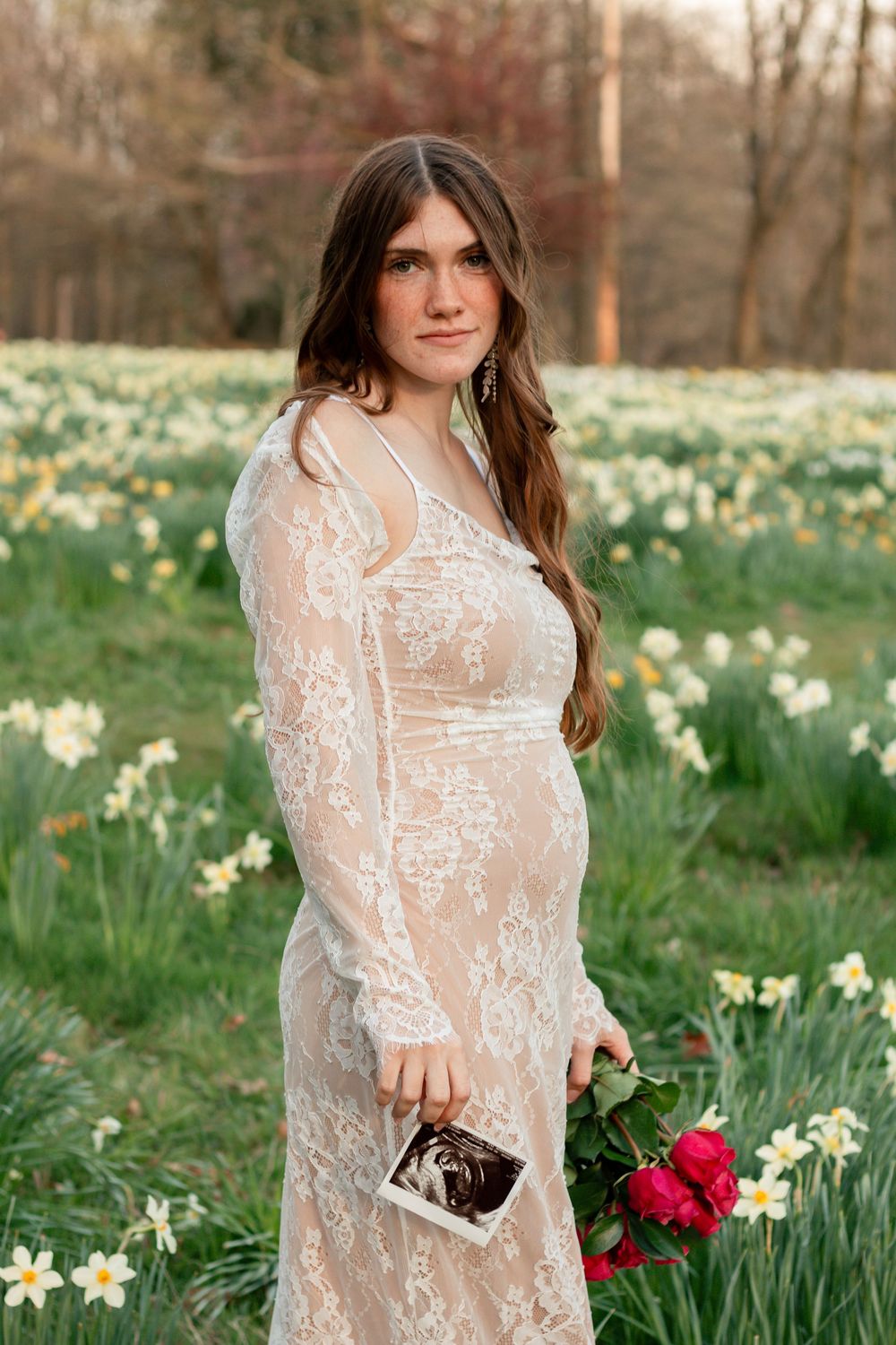 Person in white lace dress poses in field of daffodils while holding red flowers and small purse.