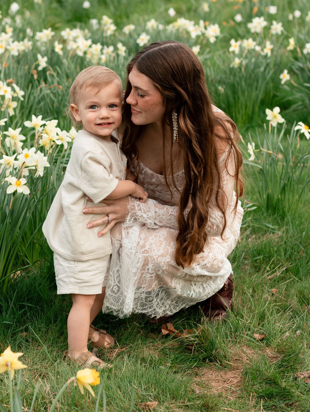 A tender moment in a spring daffodil field as a woman in lace dress embraces her small child among blooming flowers.