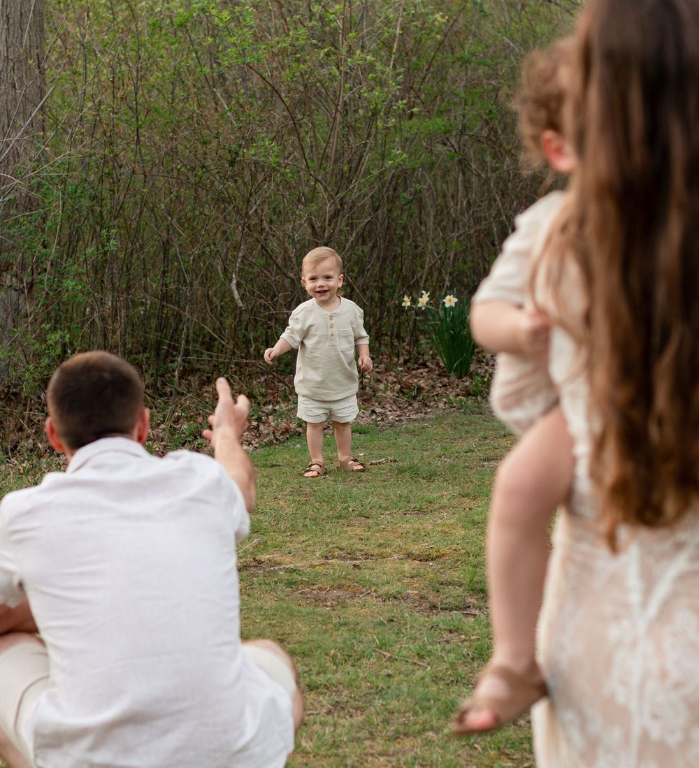Baby in neutral colored outfit takes first steps on grass while two parents watch lovingly from behind.