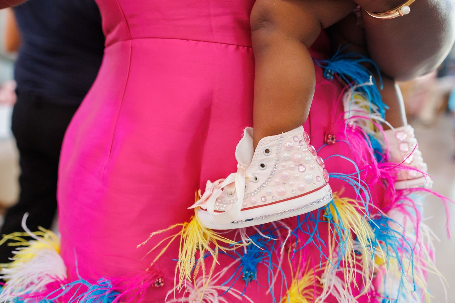 A bright pink dress with multicolored feather trim contrasts with a baby girl's white bedazzled Converse sneakers