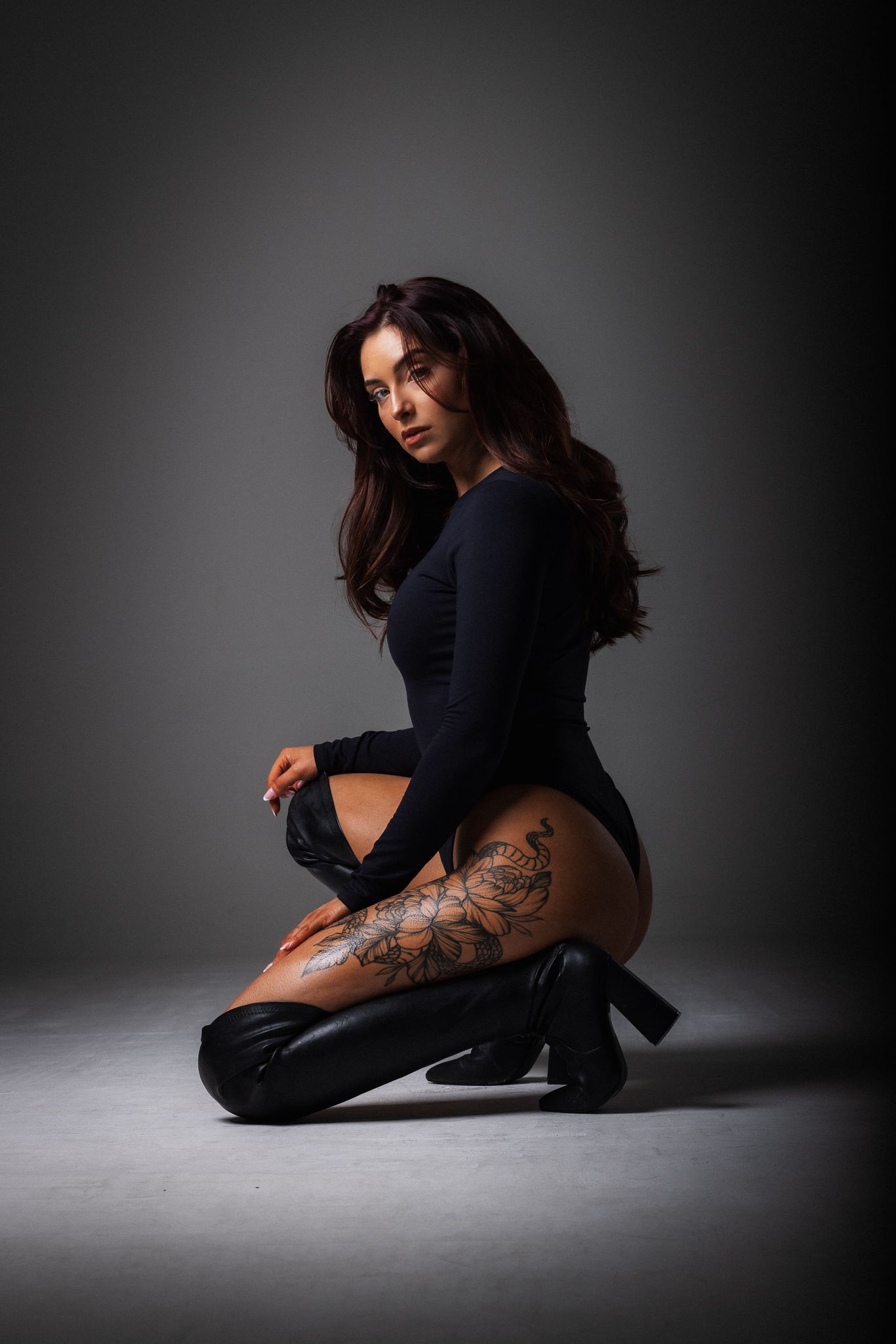 Dramatic portrait of a woman in a black bodysuit and boots posing in a dark studio setting with visible leg tattoo.