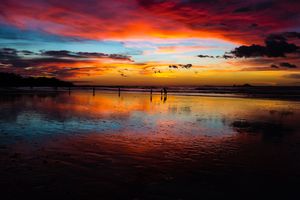 A dramatic sunset reflects vibrant orange and red colors across calm waters with silhouetted posts marking the shoreline.