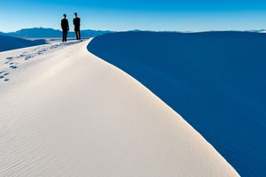 Two silhouettes stand atop a pristine white sand dune against a vibrant blue sky and mountain backdrop.