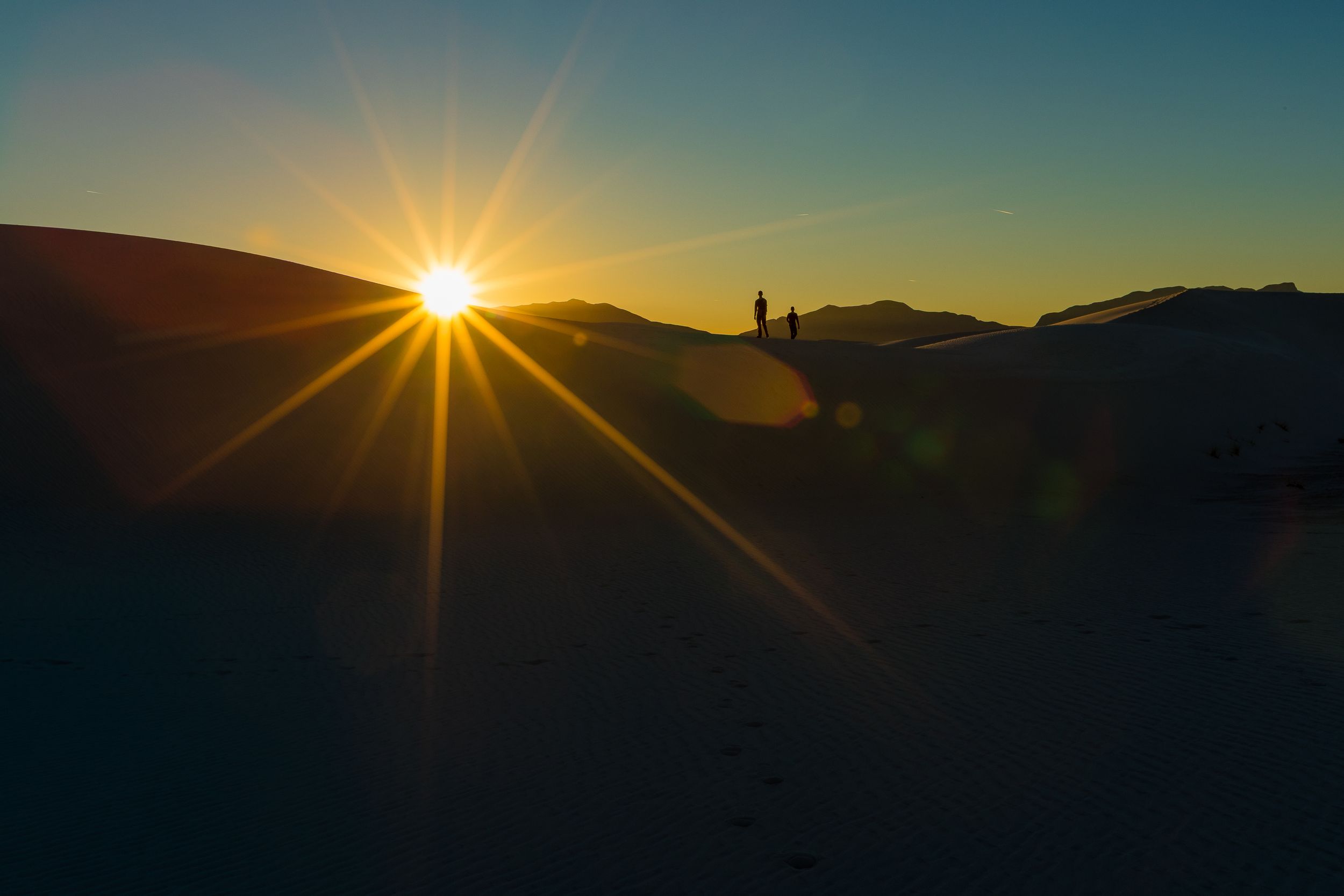 Dramatic sunset photograph showing silhouetted mountain ridges with a starburst sun effect in the orange sky.