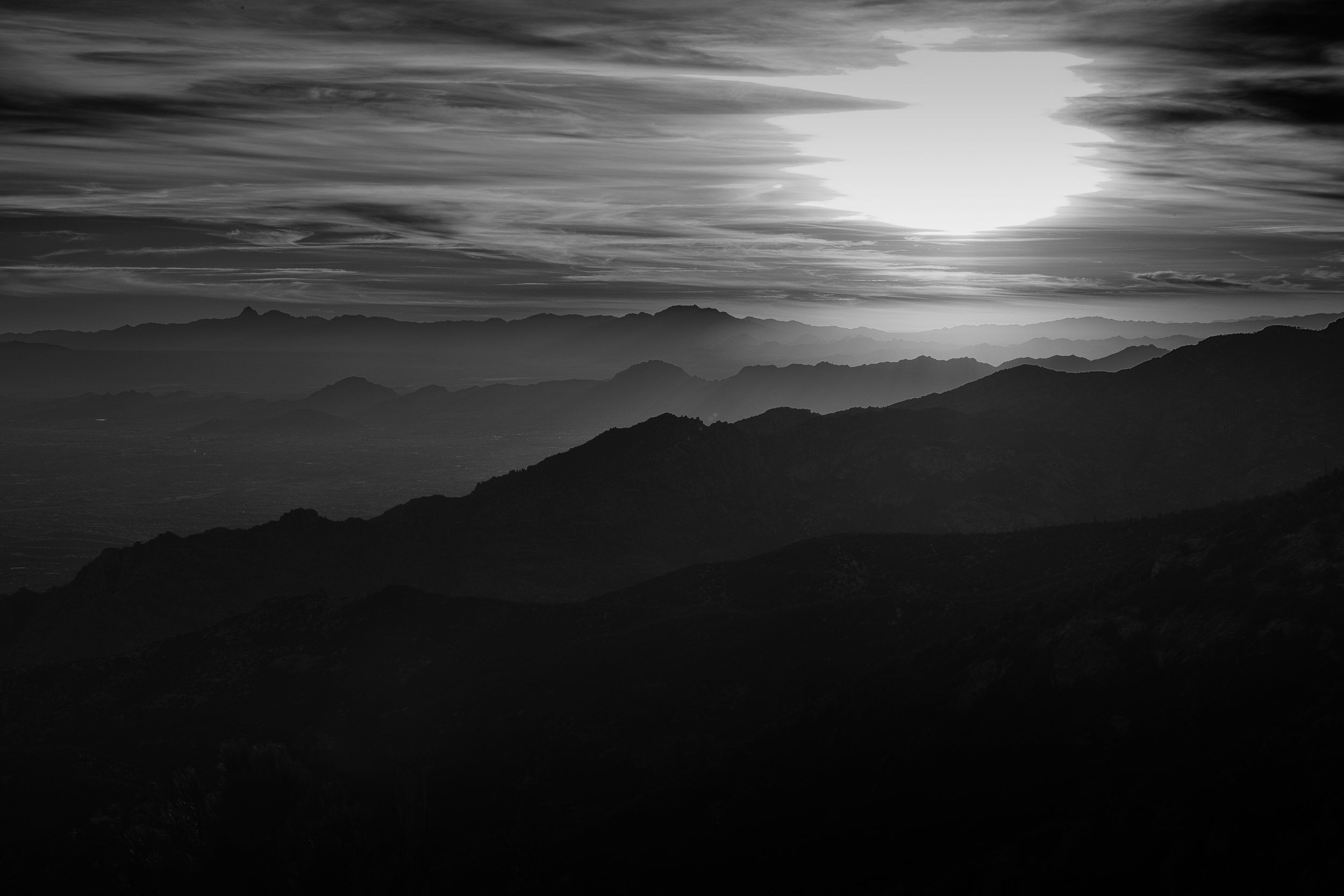 A dramatic panoramic view of mountains silhouetted against a cloudy dusk sky with a bright setting sun.