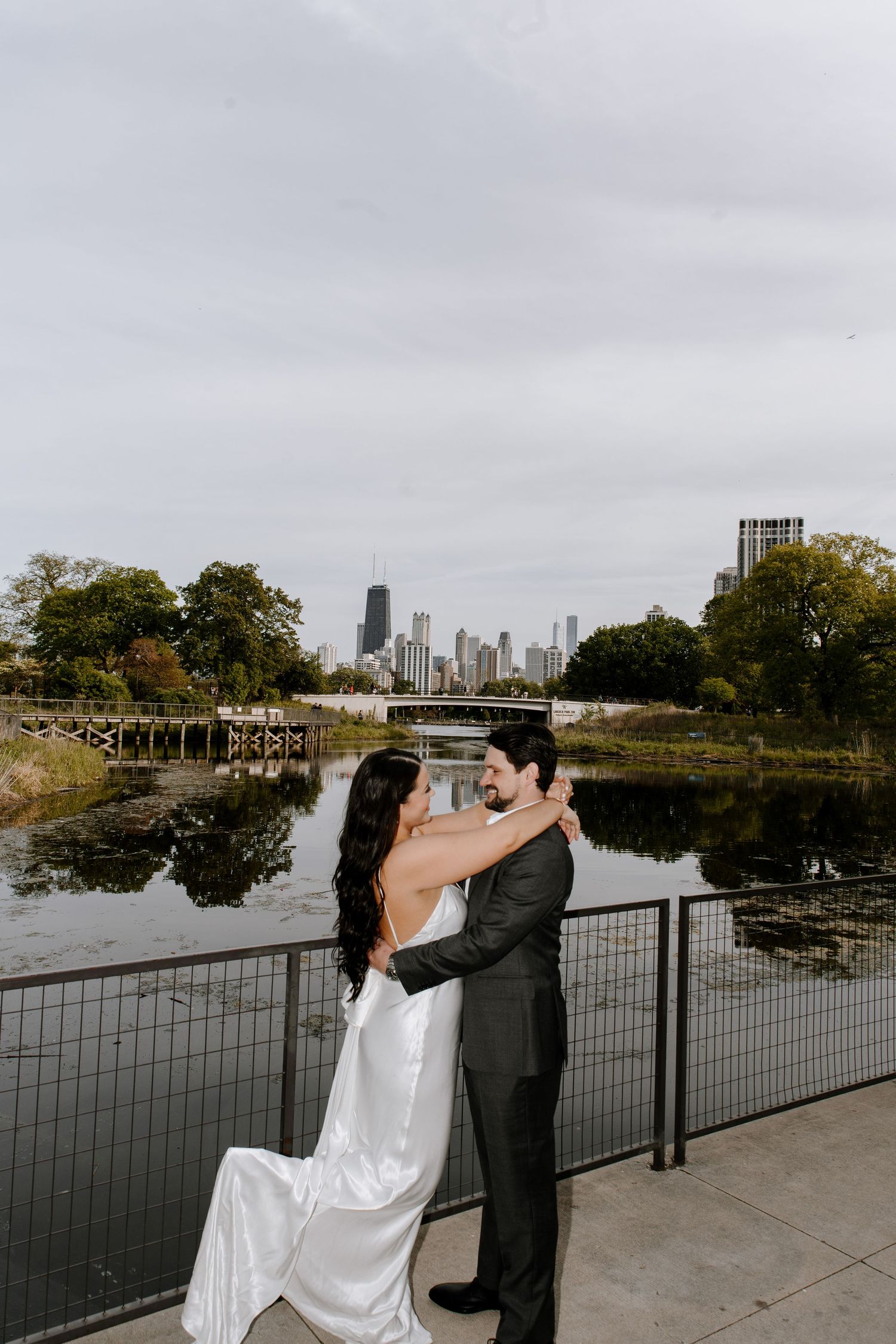 A couple stands on a bridge overlooking a pond with Chicago's iconic skyline in the background.