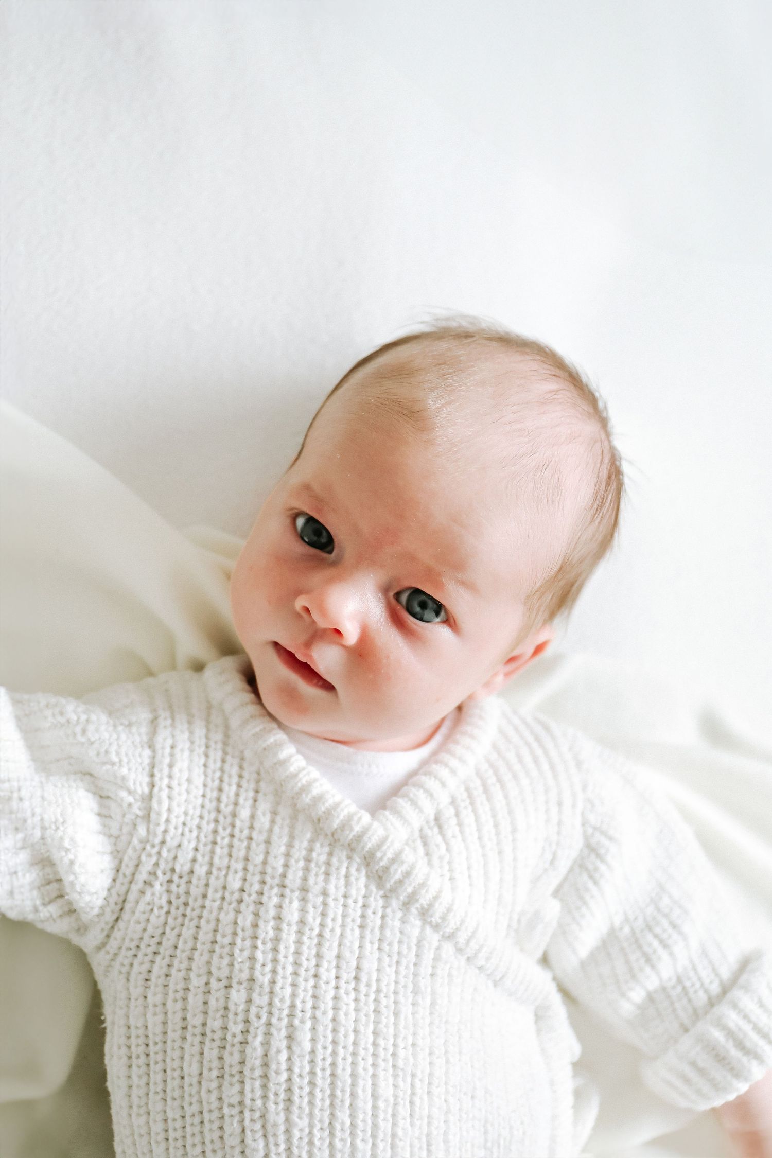 Adorable baby wearing white knit romper lying peacefully against white background, natural lighting, during newborn photoshoot in Nottingham