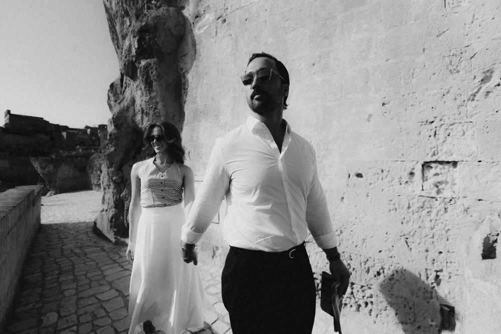 A black and white photo showing two people walking along a Mediterranean stone wall on a sunny day.