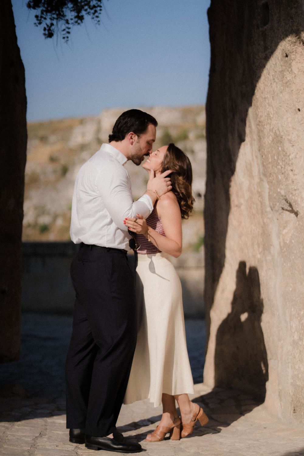 A romantic couple shares a tender moment in elegant attire against a stone wall backdrop during golden hour.