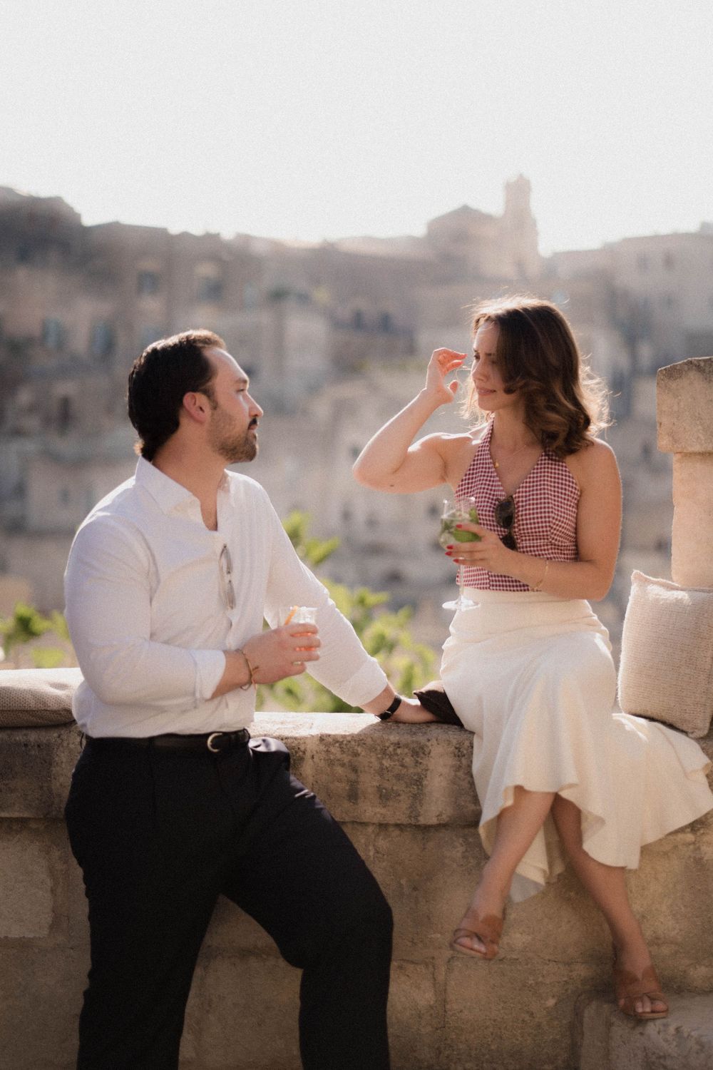 Couple enjoying drinks together on stone wall balcony overlooking historic Mediterranean cityscape at sunset.