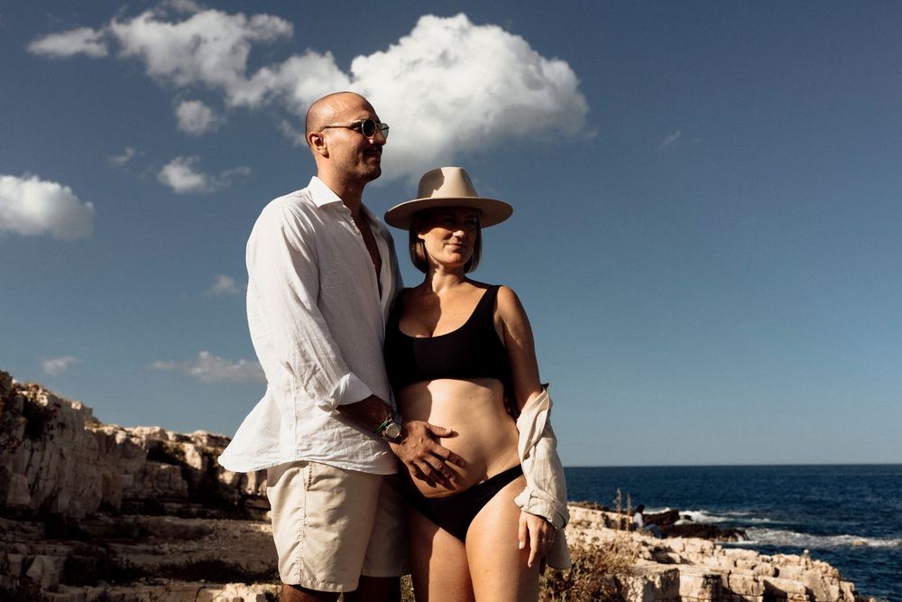 A couple poses on a rocky oceanside cliff against a blue sky with white fluffy clouds on a sunny day.