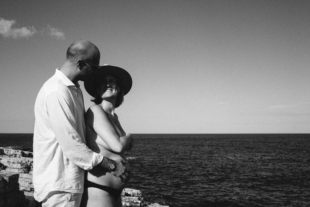 Couple embraces on cliffside overlooking Mediterranean Sea with white buildings in background.