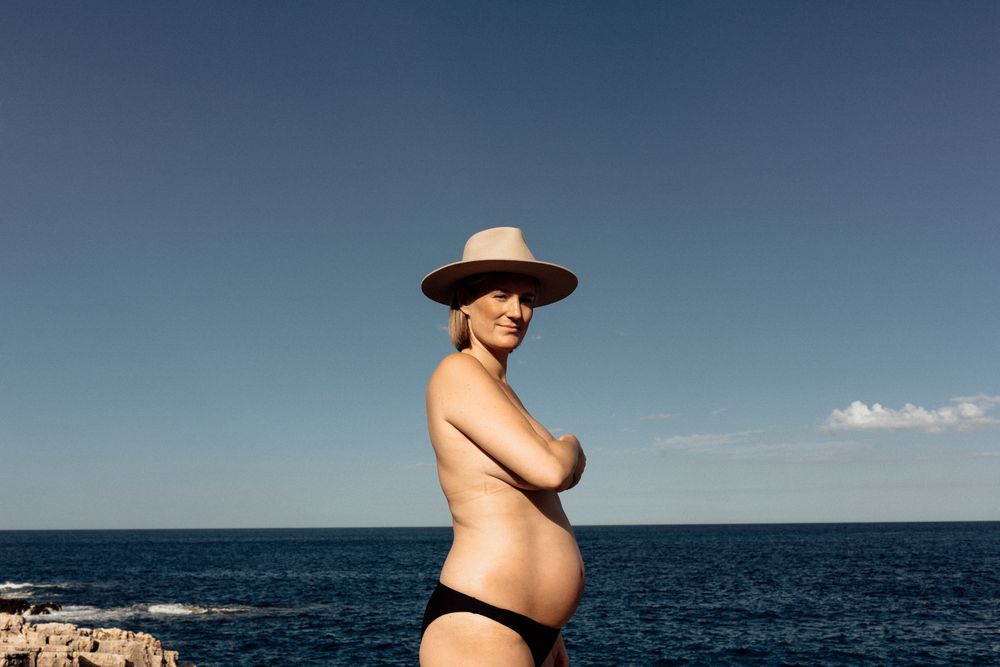 A pregnant woman in a sun hat and bikini stands against a blue ocean backdrop on a sunny day.