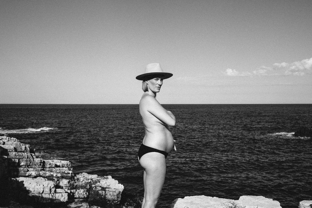 A figure in a black swimsuit and wide-brimmed hat stands elegantly on rocky coastline overlooking a vast ocean horizon.