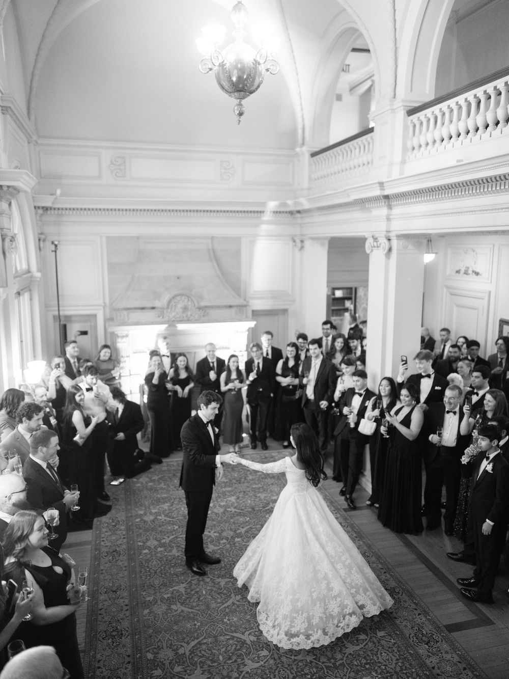 Black and white photo of bride and groom sharing first dance in elegant ballroom surrounded by guests.