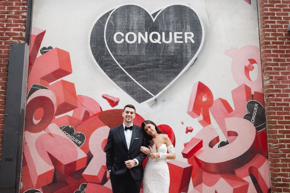 Wedding couple poses joyfully by the bold "Conquer" mural near Lilah in Philadelphia — a perfect blend of urban art and romance.