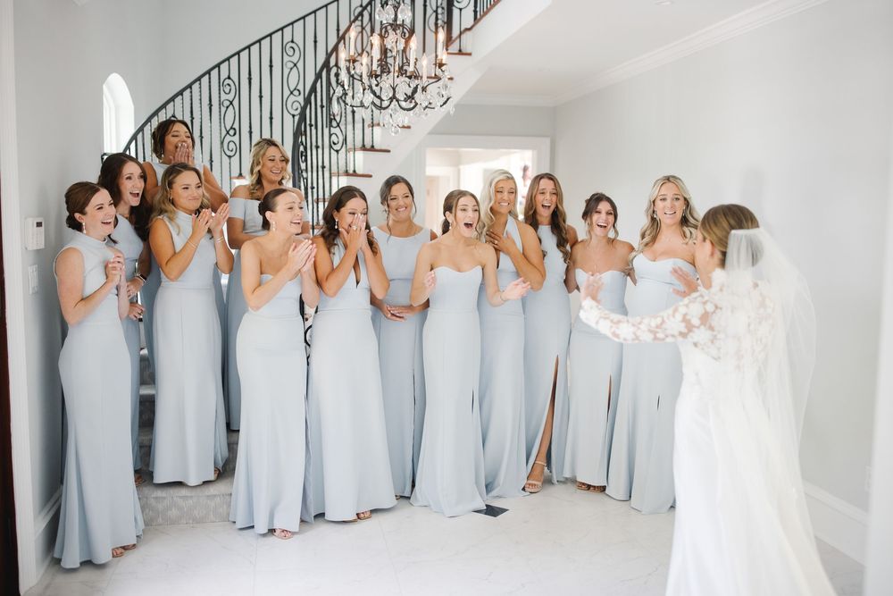 Bride reveals her wedding dress to an excited bridal party in soft blue gowns—perfect getting-ready moment full of joy and elegance.