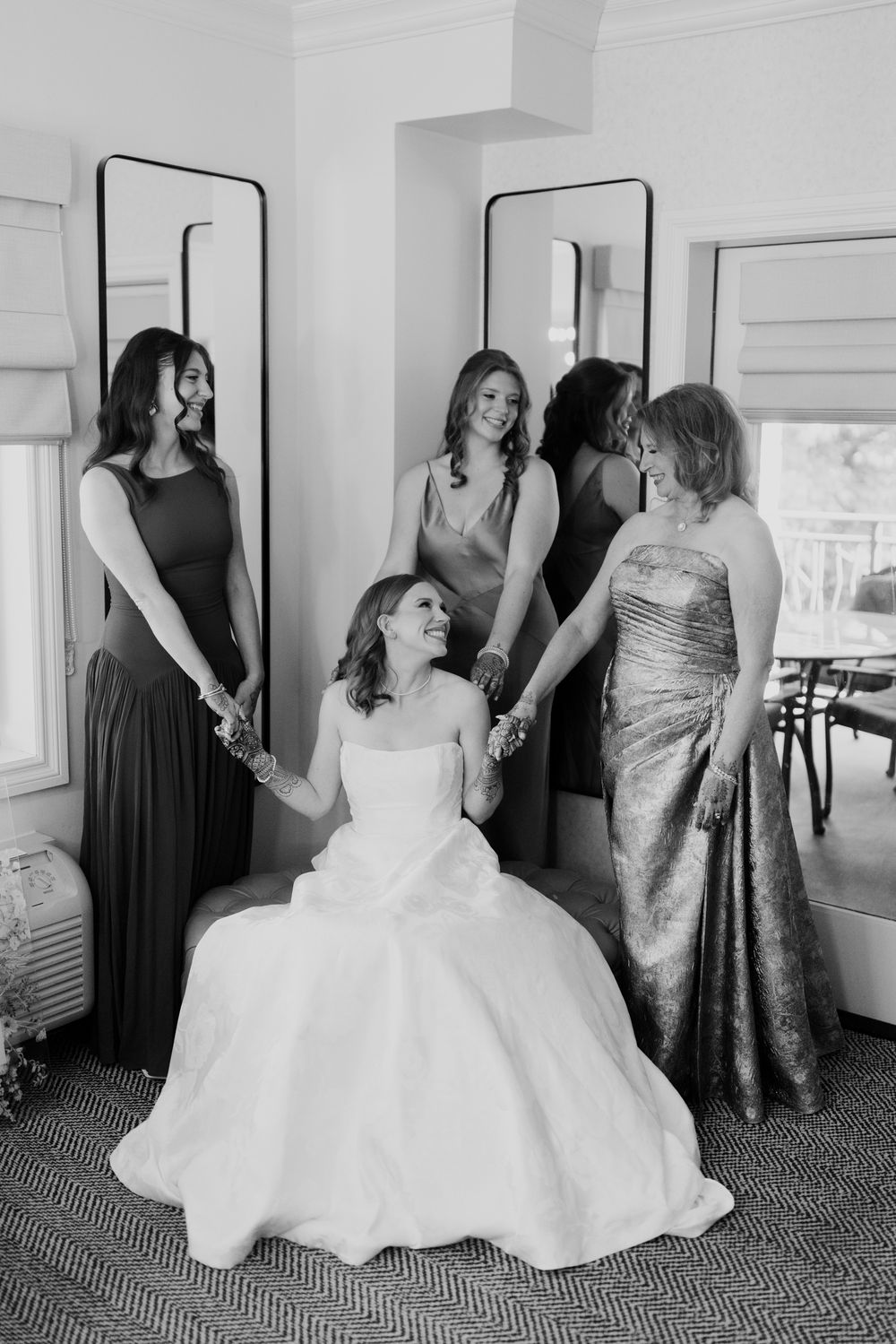 Bride seated in wedding gown holding hands with bridesmaids and mother in elegant black and white getting ready photo.