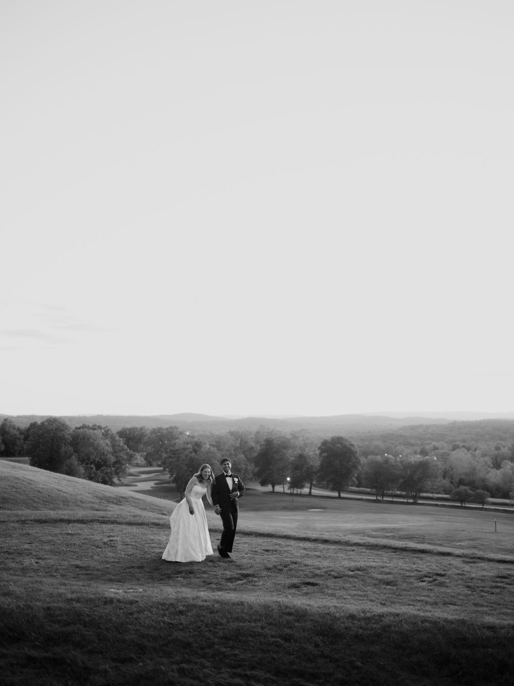 Black and white photo of a bride and groom standing on a hillside overlooking a scenic landscape at dusk.