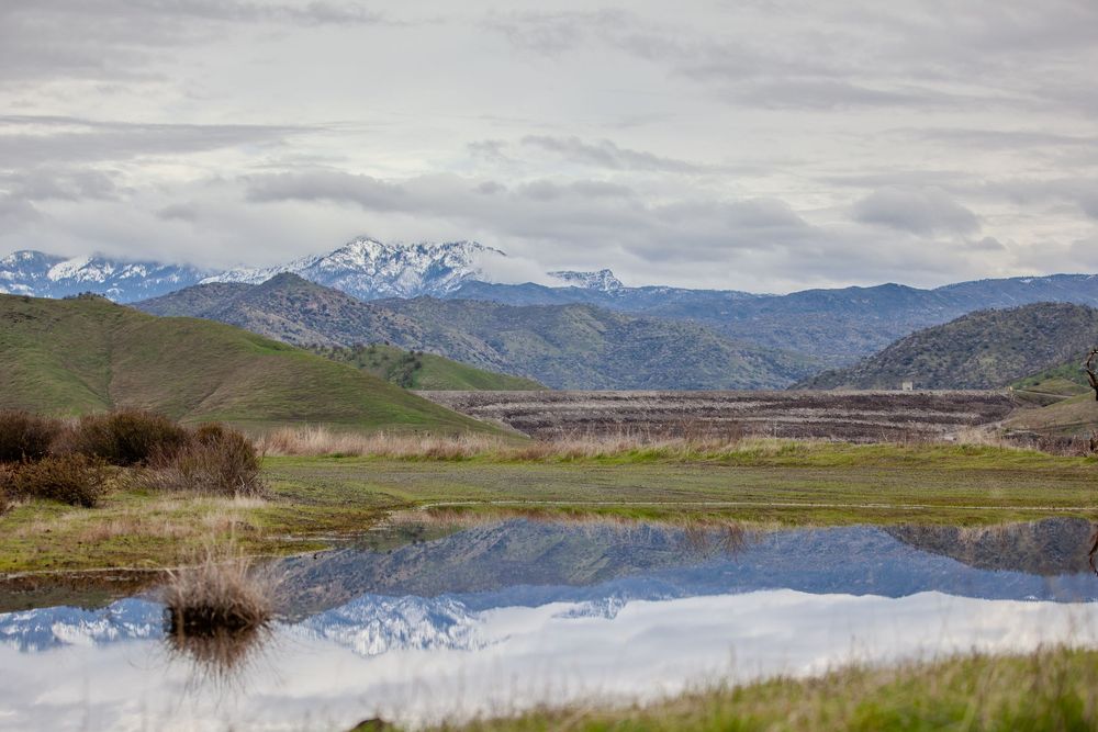 Snow-capped mountains reflect in a calm wetland pool under cloudy skies.