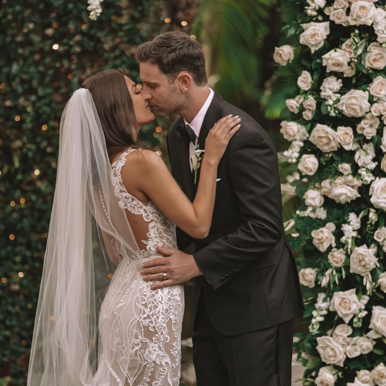 A romantic wedding moment captured against a backdrop of white roses and greenery at an outdoor ceremony.