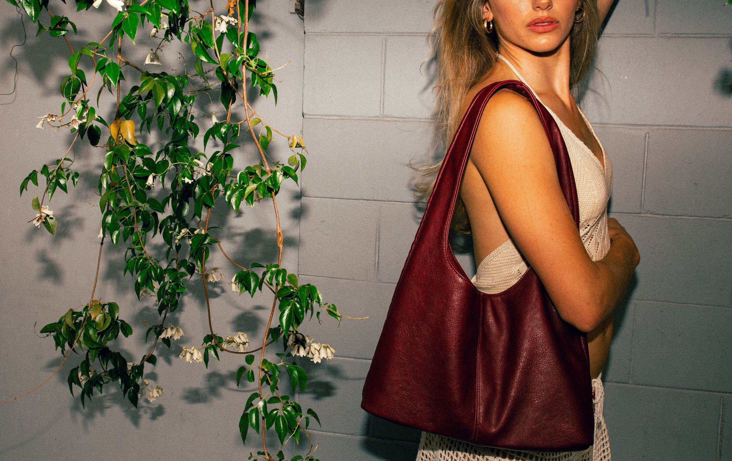 Side view of someone wearing a burgundy tote bag and tank top standing near green ivy on a concrete wall.