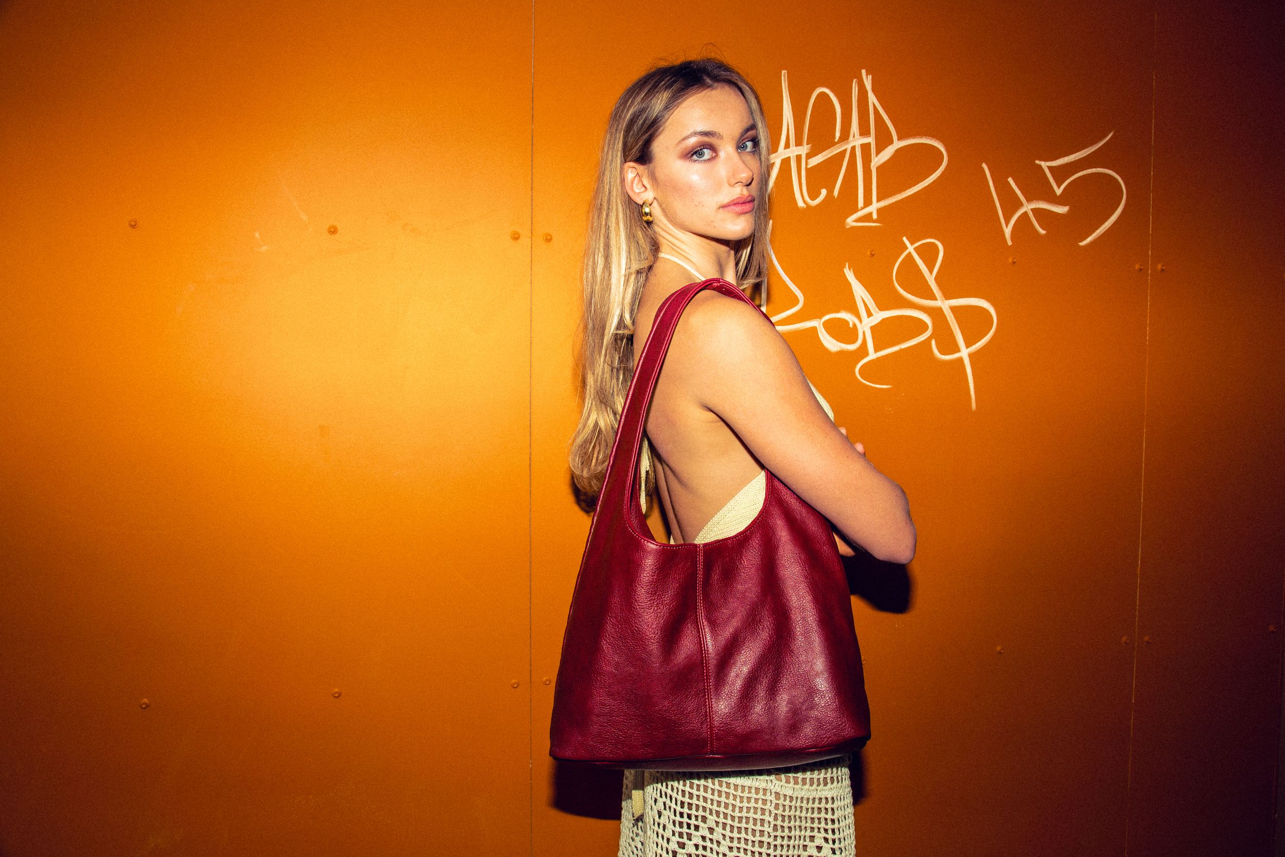 A person wearing a burgundy tank top poses in front of an orange-lit wall with graffiti.
