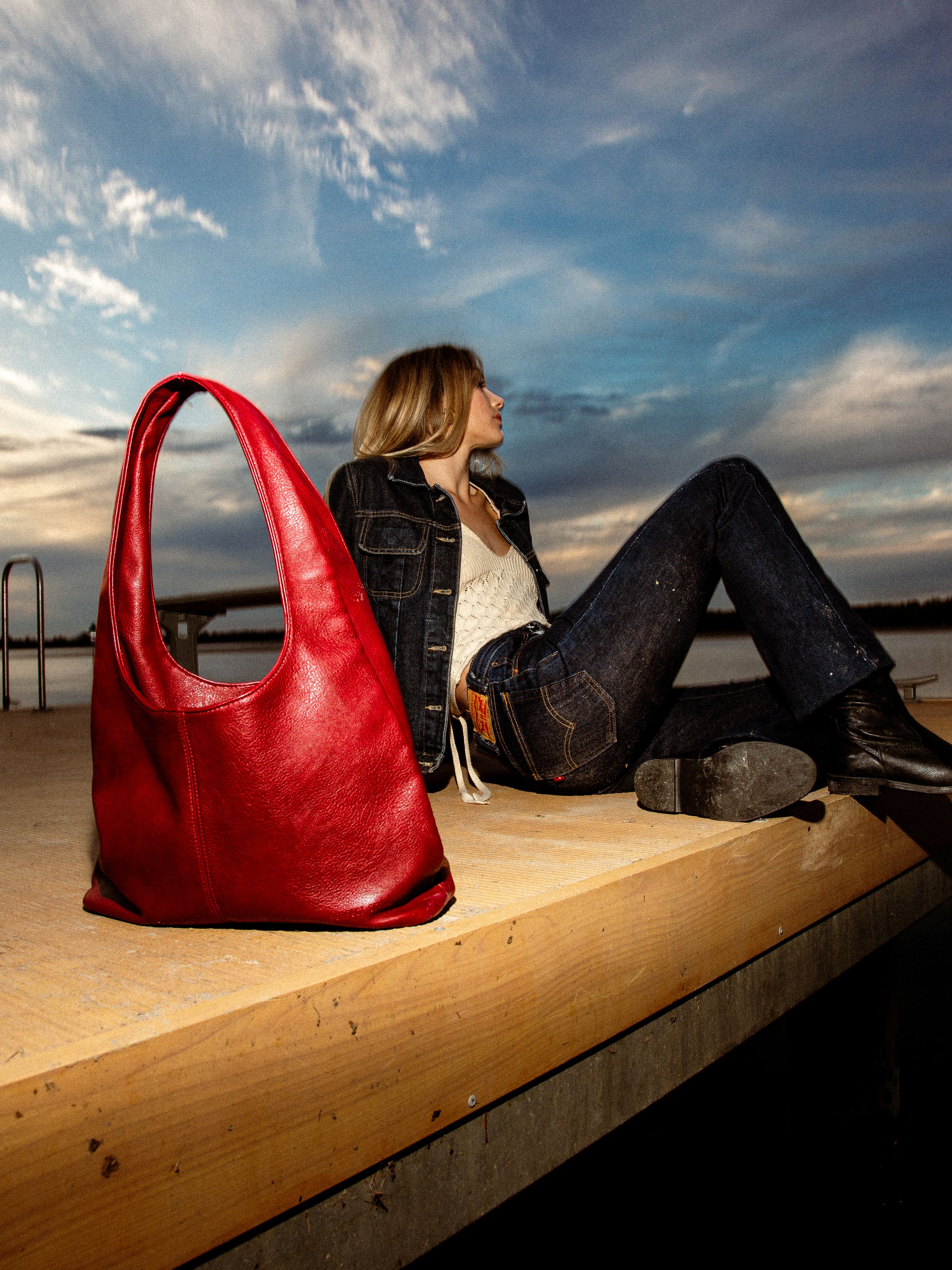 Stylish red leather hobo bag sits beside relaxed figure on wooden dock at sunset with dramatic cloudy sky.