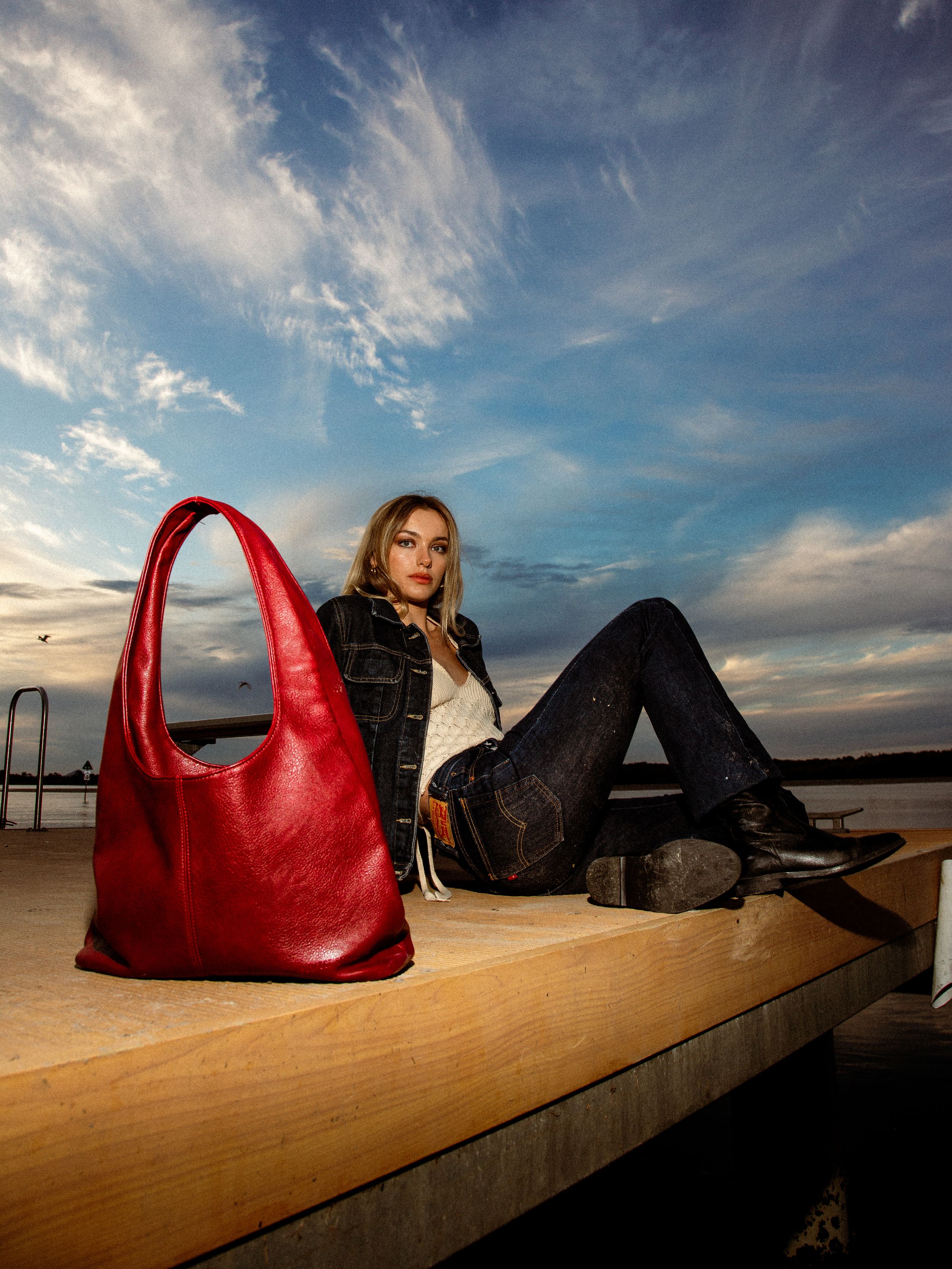 A bright red leather handbag sits on a wooden platform against a dramatic sunset sky and beach background.