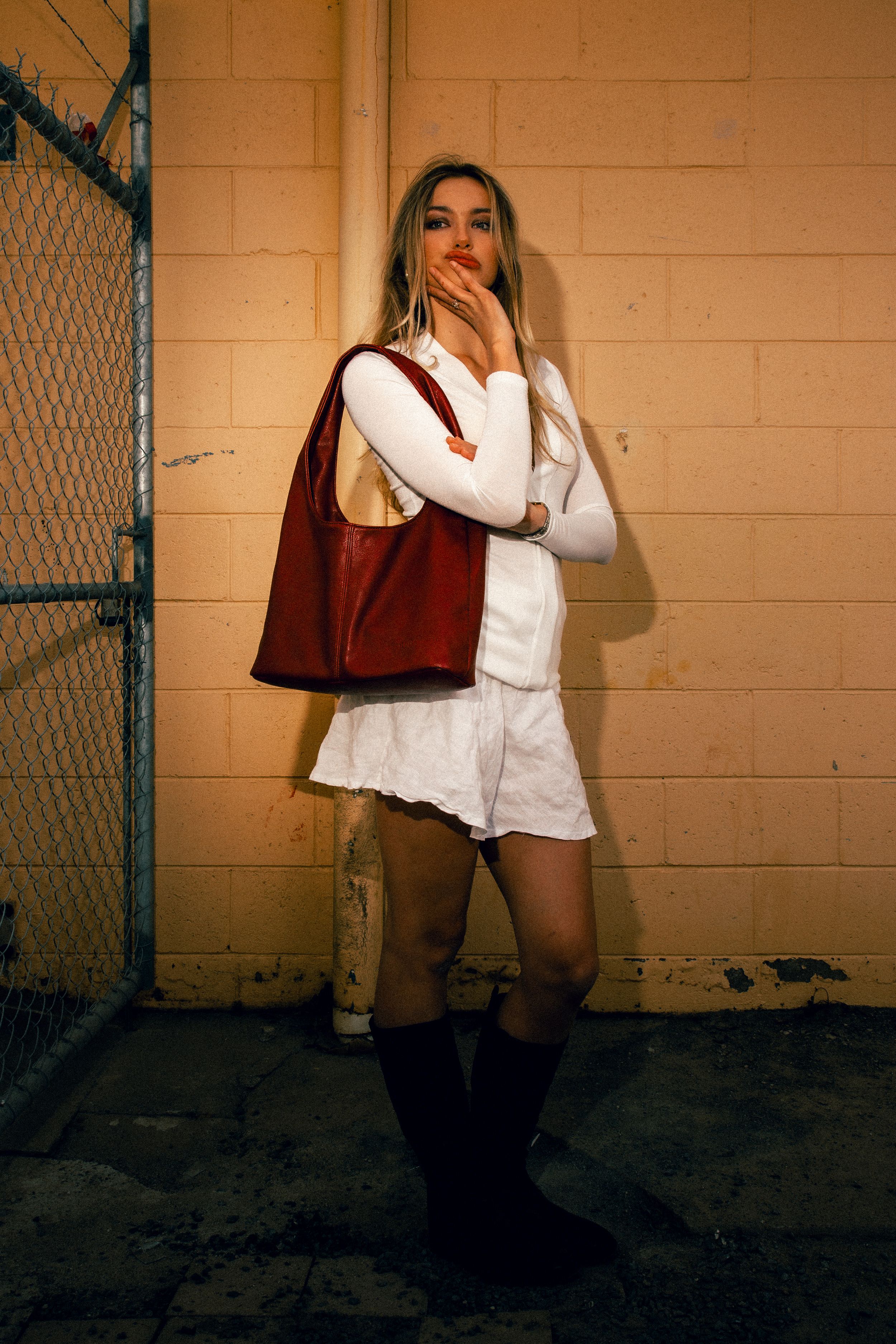 A person in a white dress poses against a wall while carrying a brown leather tote bag and wearing tall black boots.