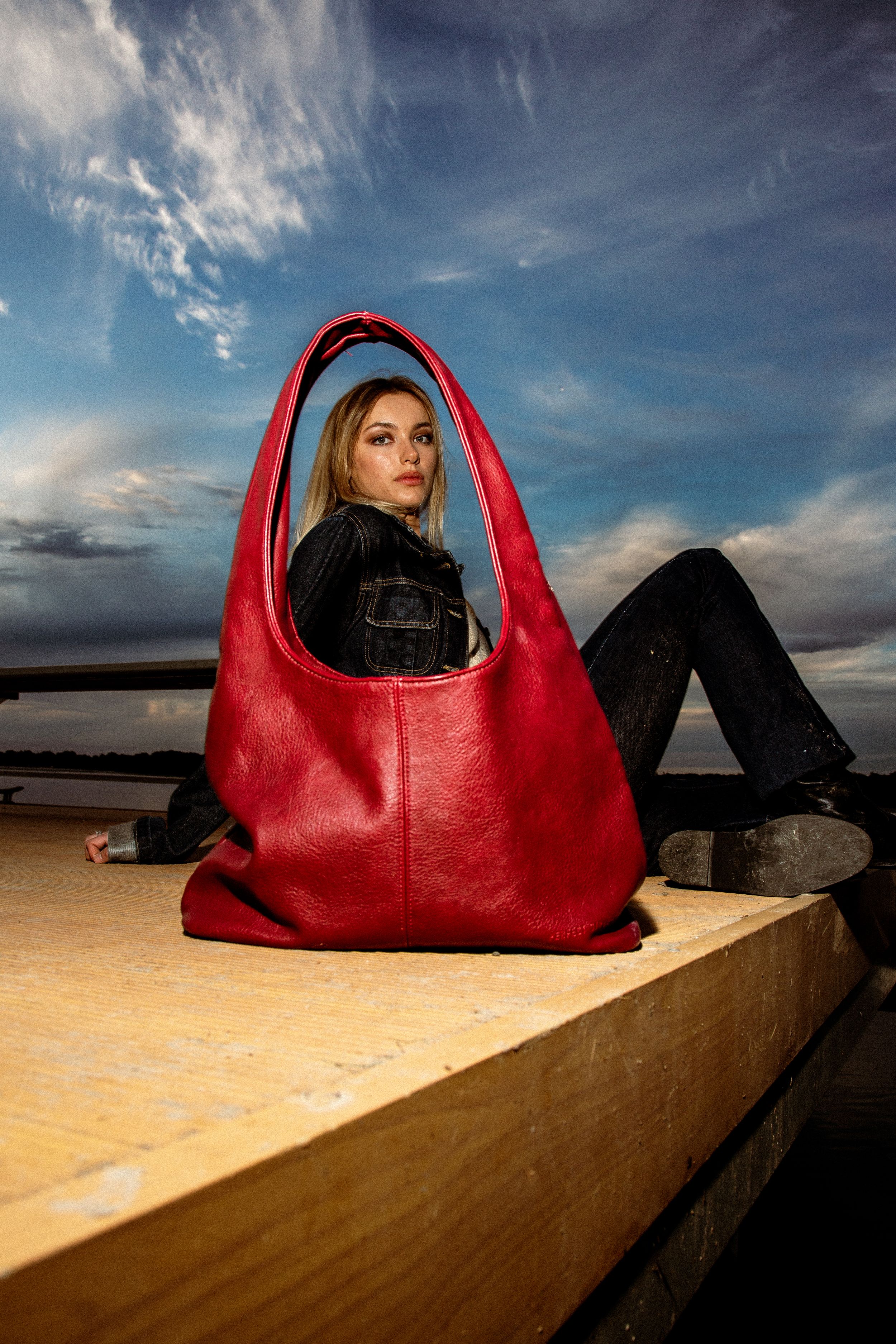 A vibrant red hobo bag is displayed on a wooden surface with moody clouds and beach scenery in the background.