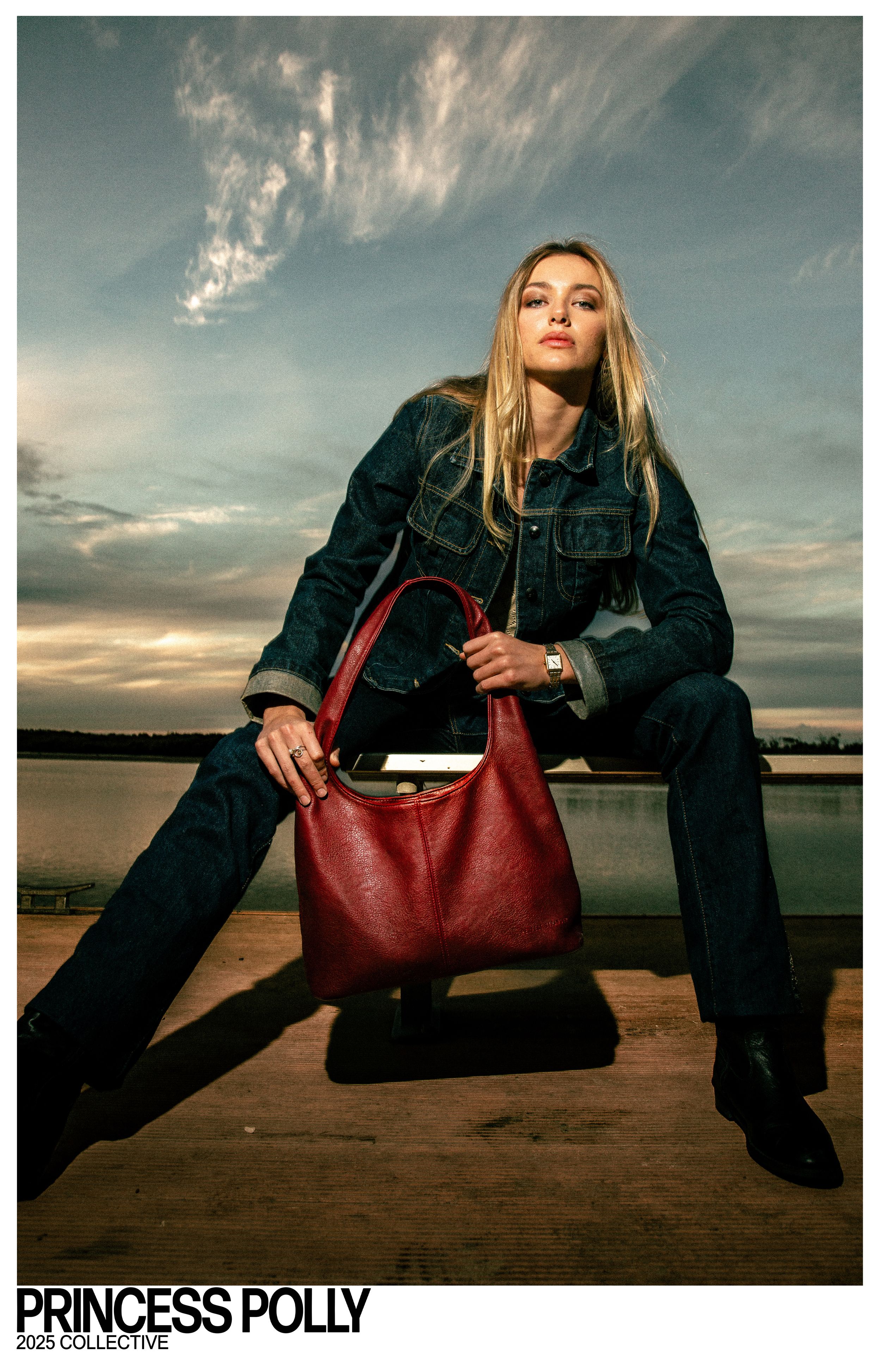 Fashion photography at sunset shows model in denim outfit with red leather handbag sitting on wooden dock by lake.