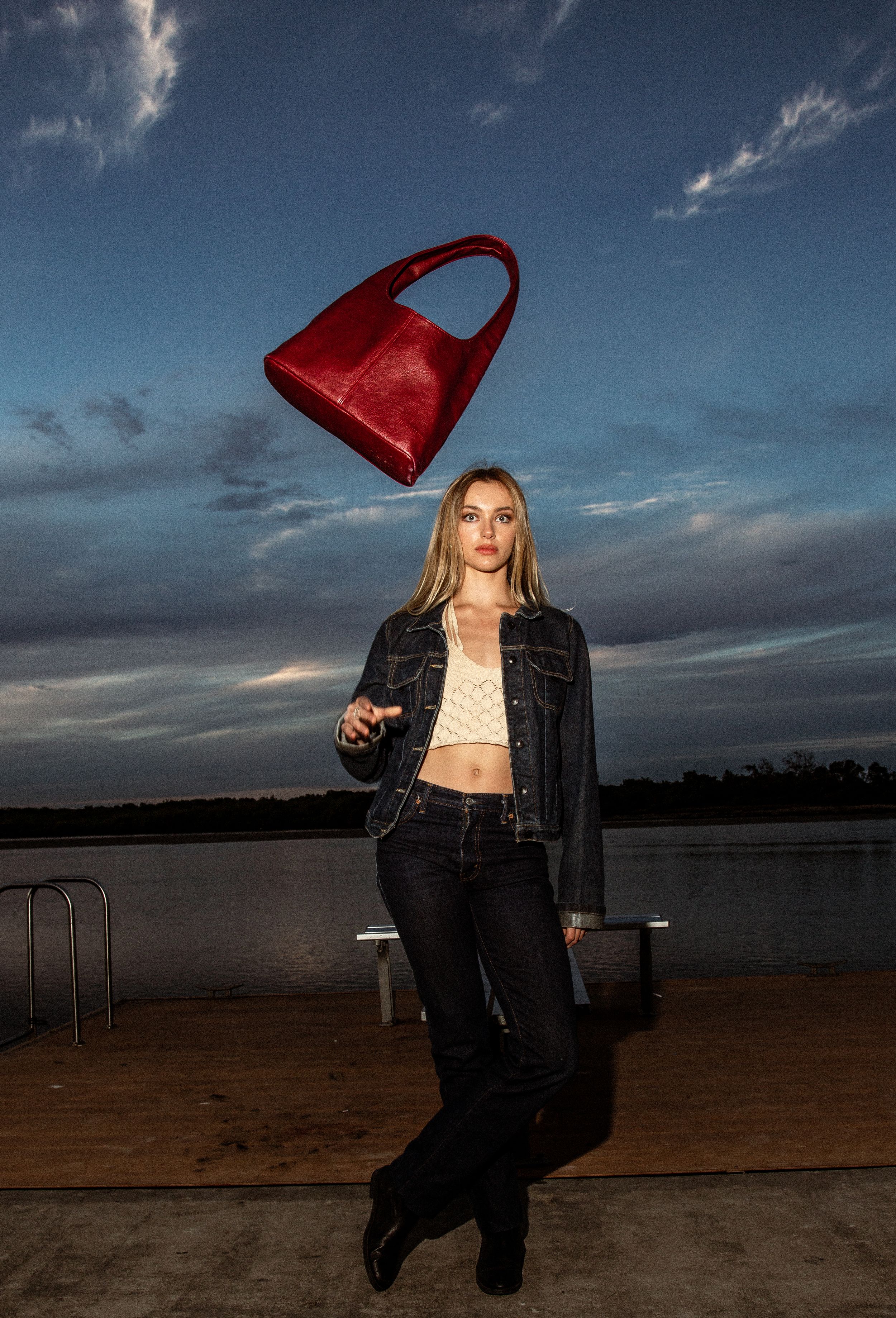 Fashion model poses outdoors at sunset with a red handbag against dramatic skyline and water backdrop.
